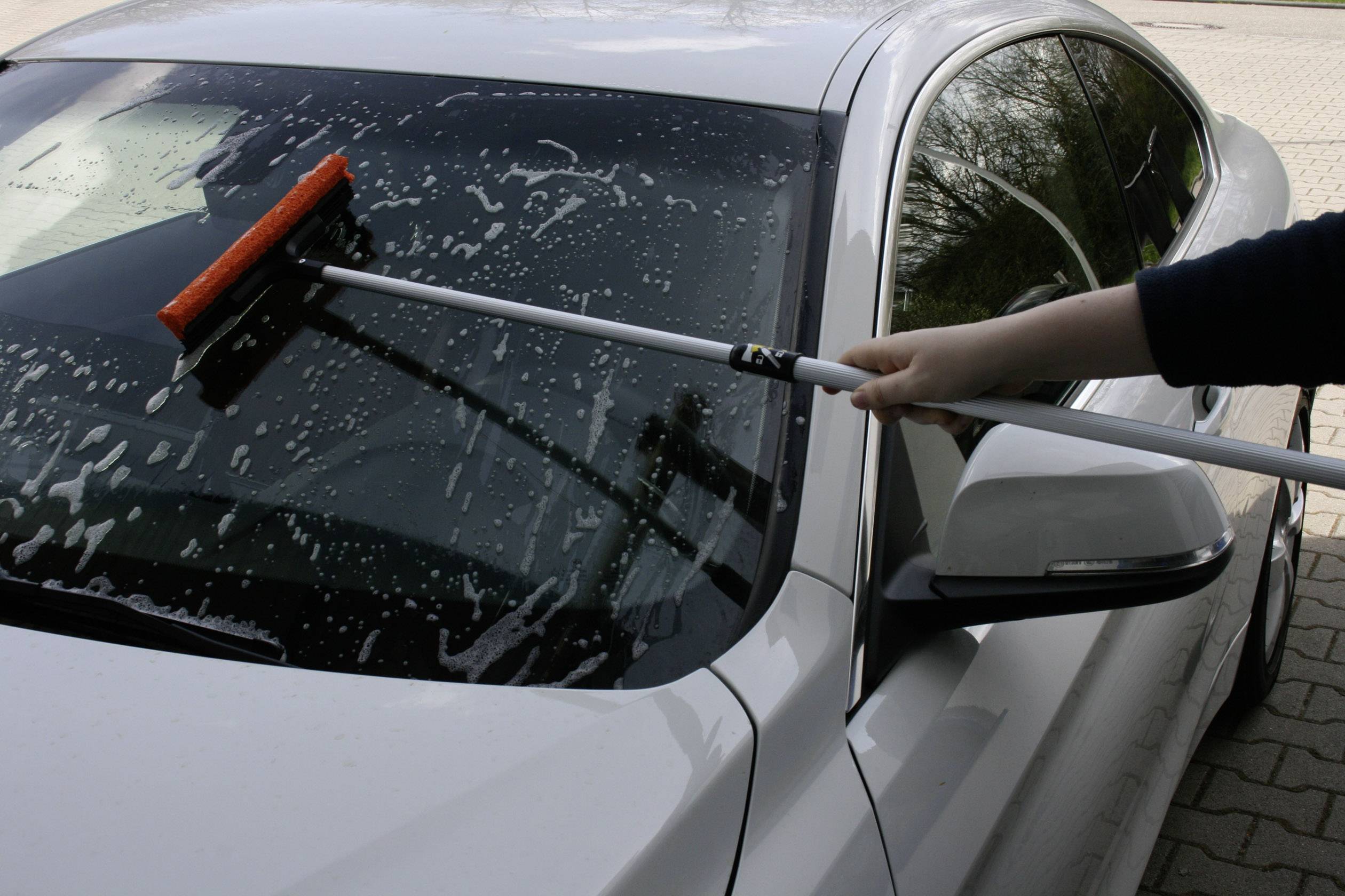 Een persoon reinigt de voorruit van een witte auto met een lange bezem op een parkeerplaats.