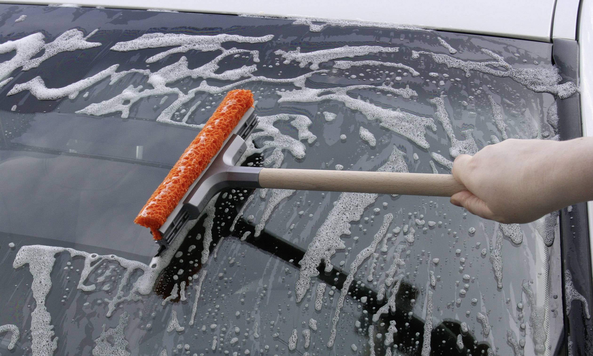 Een hand reinigt de voorruit van een auto met een raamwisser en zeepwater. De achtergrond toont delen van het voertuig.
