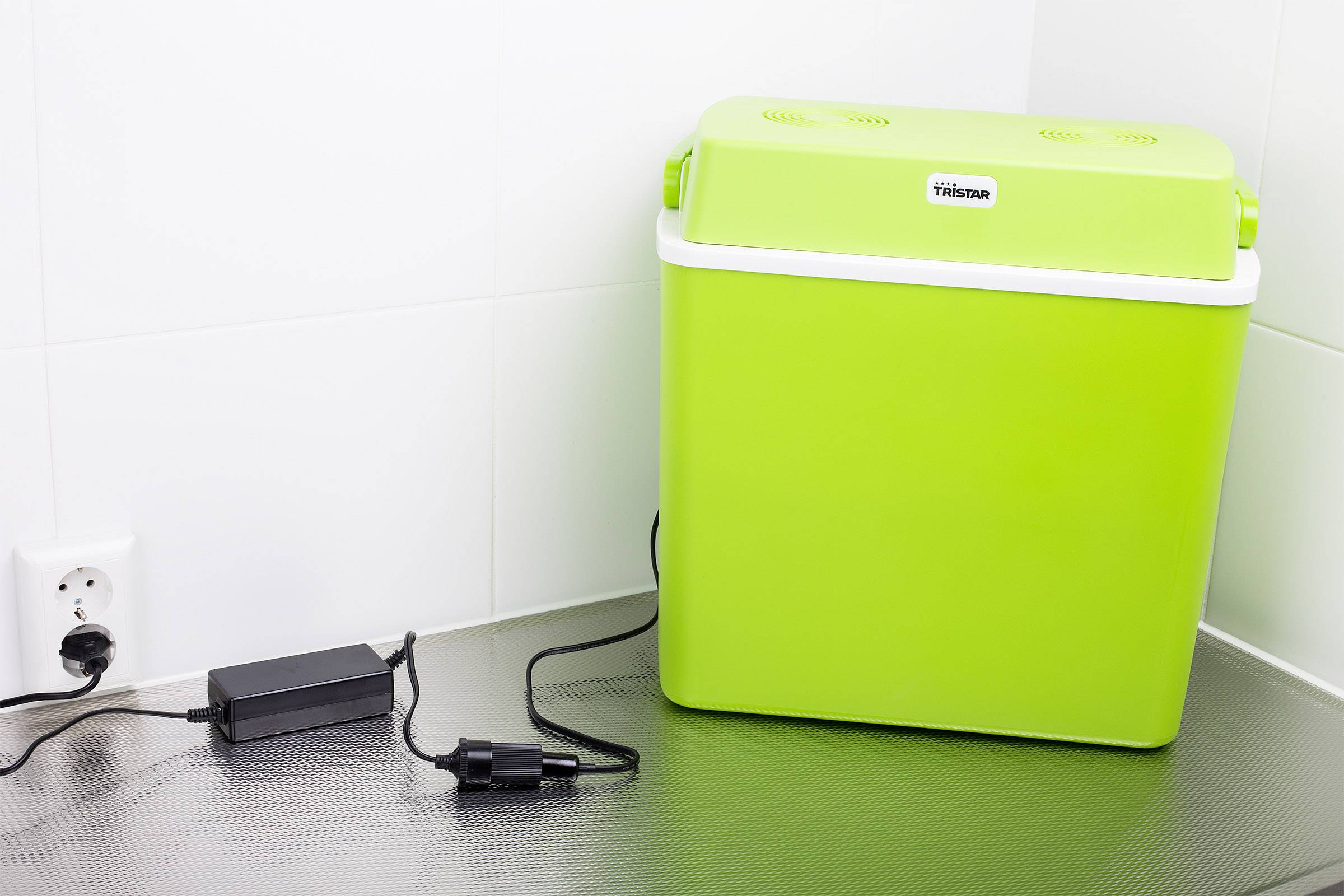 Kleine groene koelbox op een metalen oppervlak naast een stopcontact. Een zwarte voedingsadapter is aangesloten, waarvan het snoer naar het stopcontact loopt.
