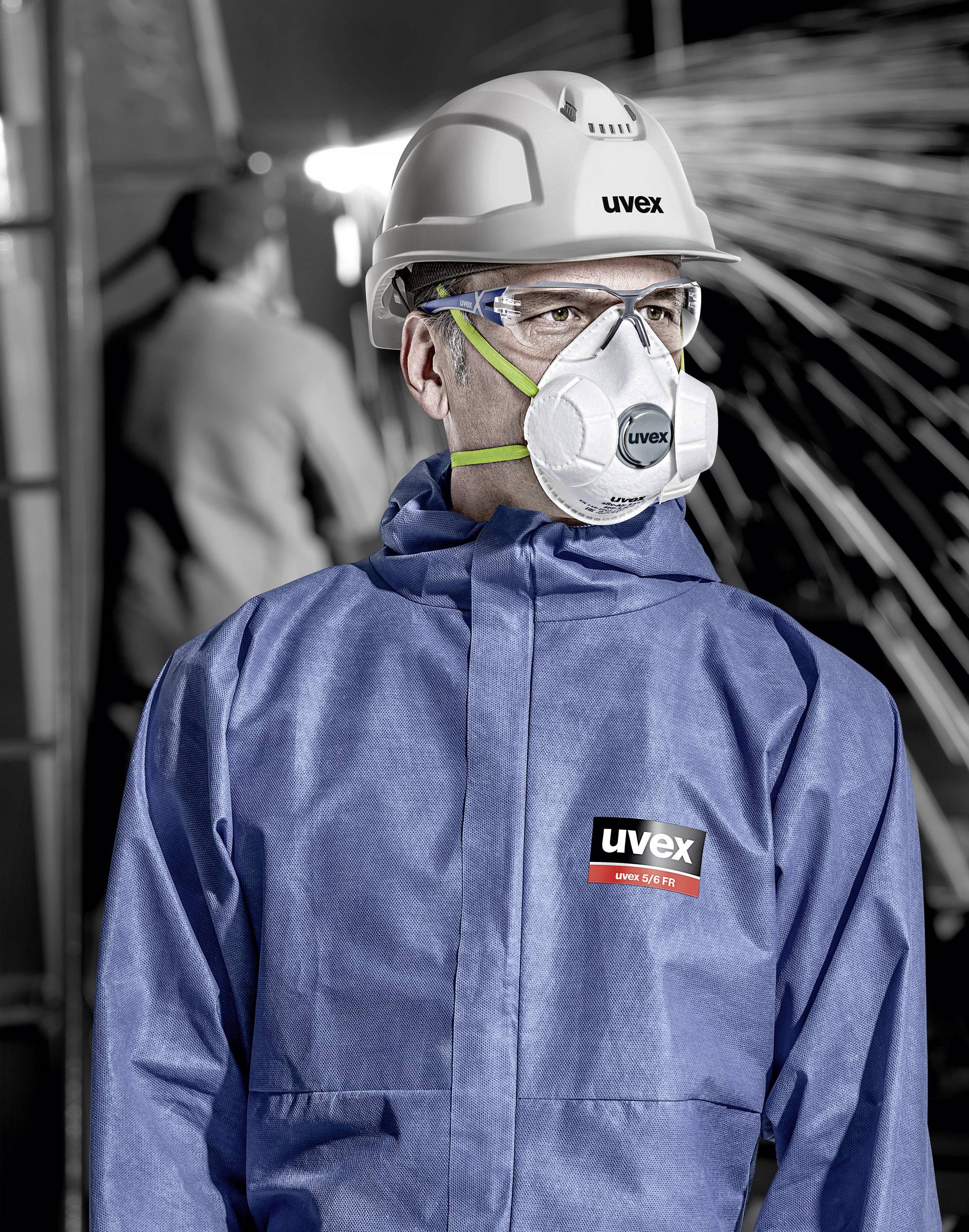 Persoon in blauwe werkkleding, met helm en veiligheidsmasker, staat in een industriële omgeving met vonken op de achtergrond.