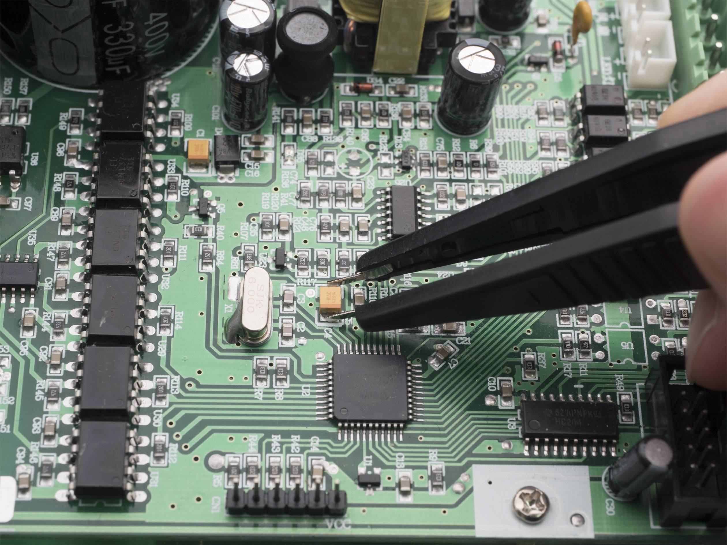 Close-up van een hand die met een pincet een klein elektronisch onderdeel op een groene printplaat plaatst.