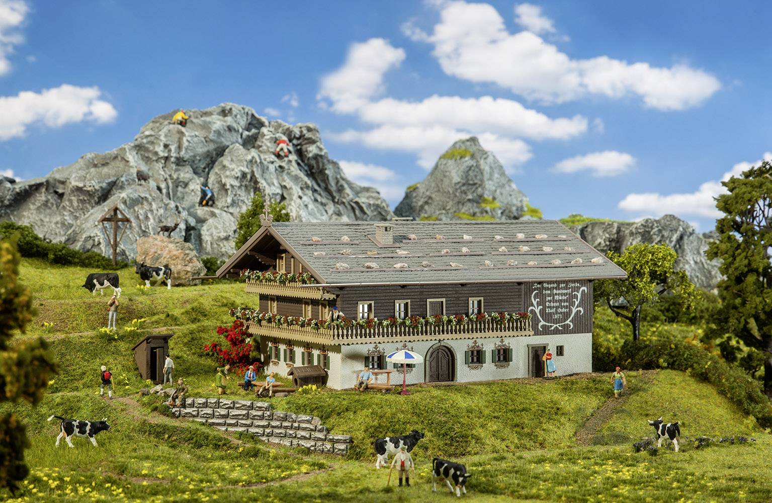 Een miniatuur boerenhuis in een berglandschap met koeien, een wandelaar en een blauwe lucht op de achtergrond.