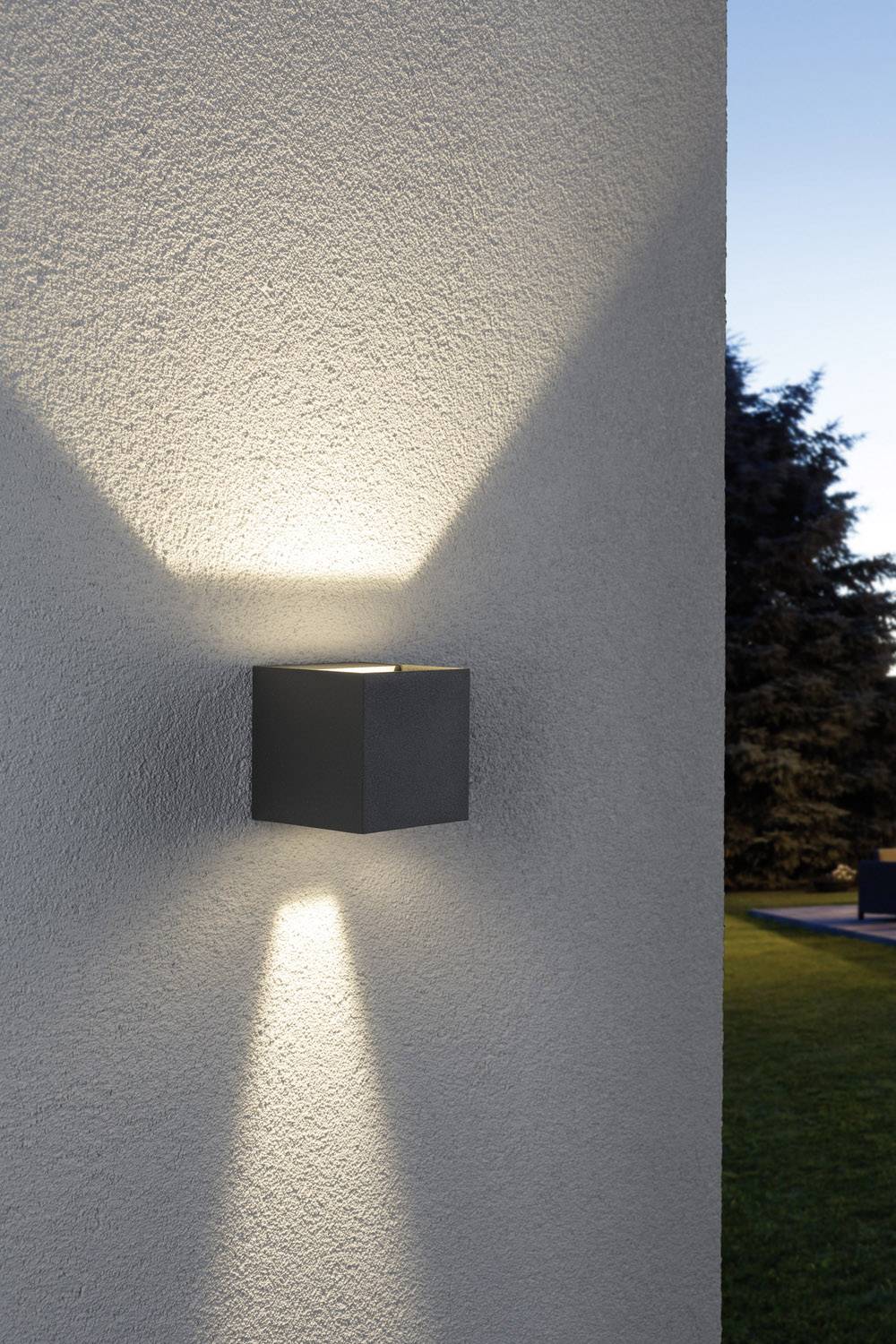 Wandlamp aan een witte muur die warm licht uitstraalt naar boven en beneden, verlicht subtiel de buitenruimte bij het invallen van de schemering.