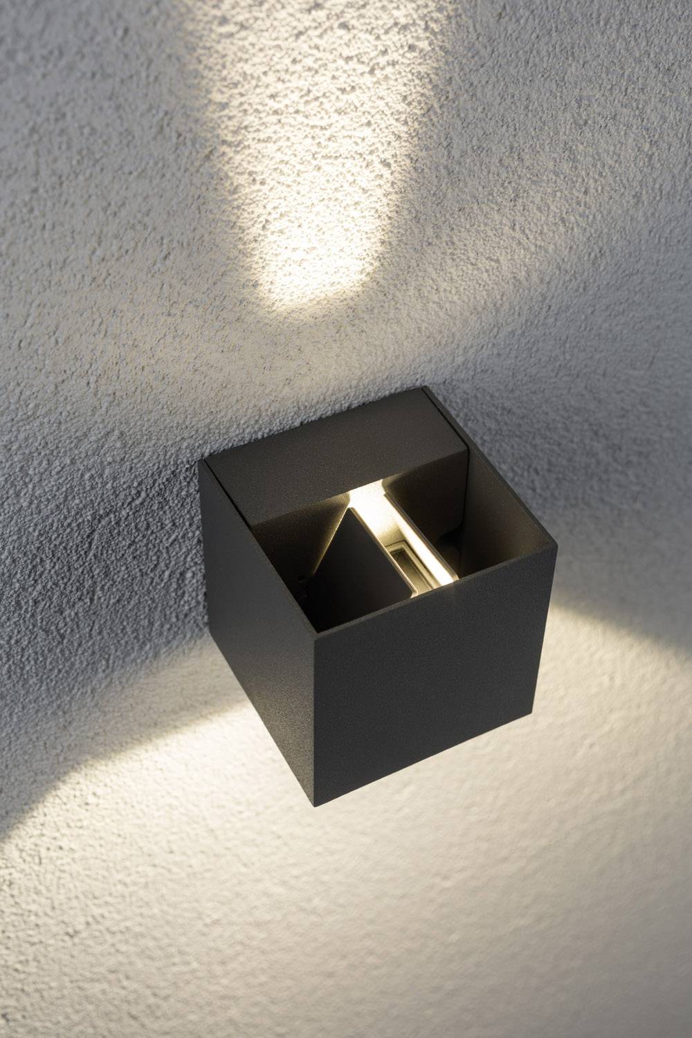 Wandlamp van een zwarte metalen kubus werpt licht naar boven en beneden langs een witte muur, creëert elegante verlichtingseffecten.