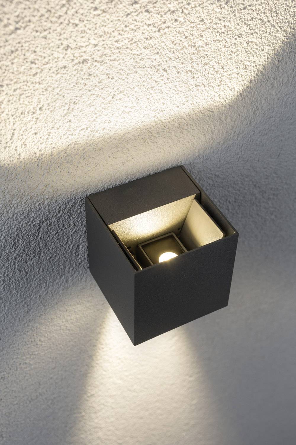 Wandlamp in modern, zwart kubusontwerp straalt omhoog en omlaag tegen een gestructureerde muur.