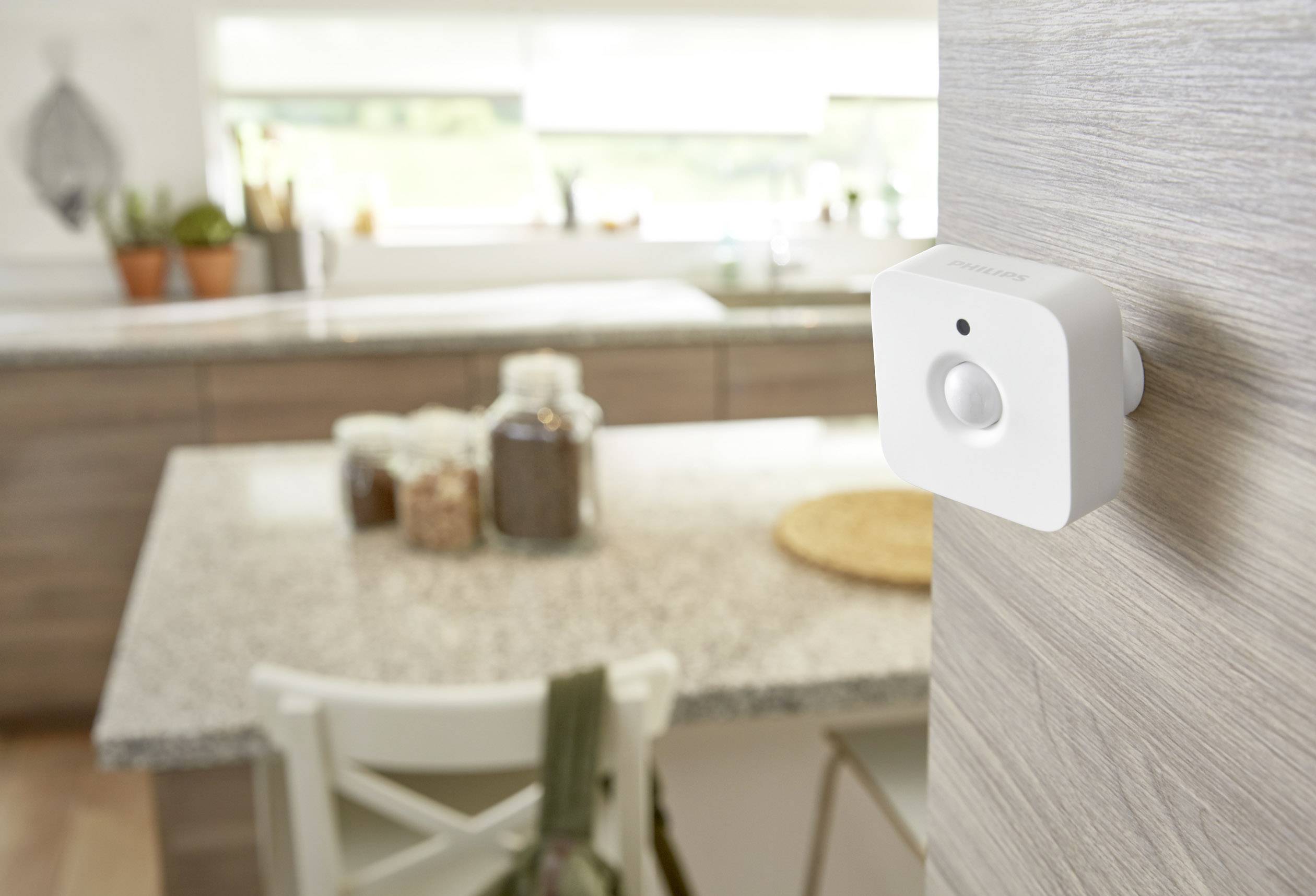 Een bewegingssensor is gemonteerd aan een houten muur in een moderne keuken. Op de achtergrond staat een tafel met voorraadcontainers.