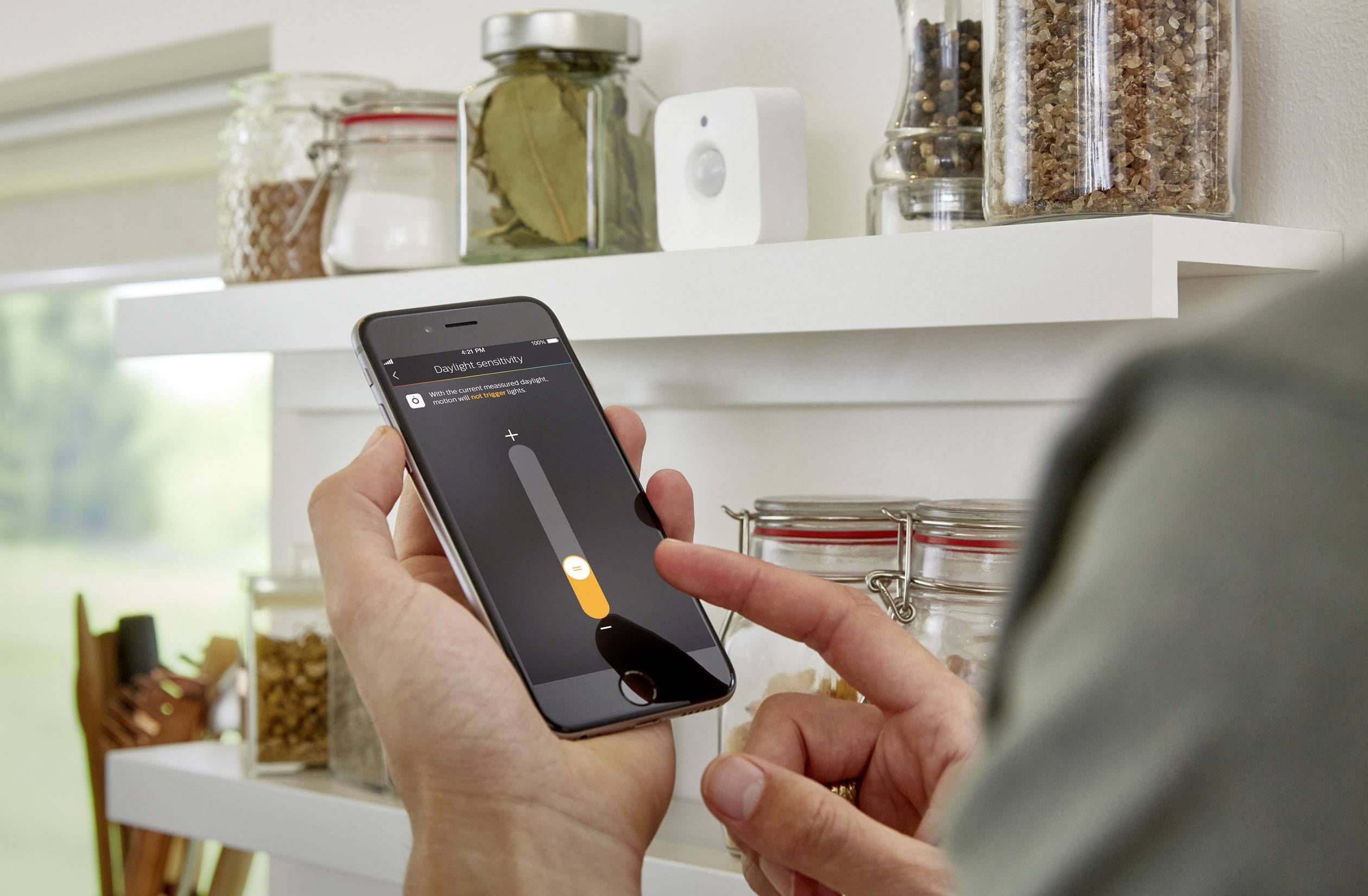 Een persoon bedient een smartphone met een app voor het beheren van een intelligent verlichtingssysteem. In de achtergrond staat een rek met kruiden.