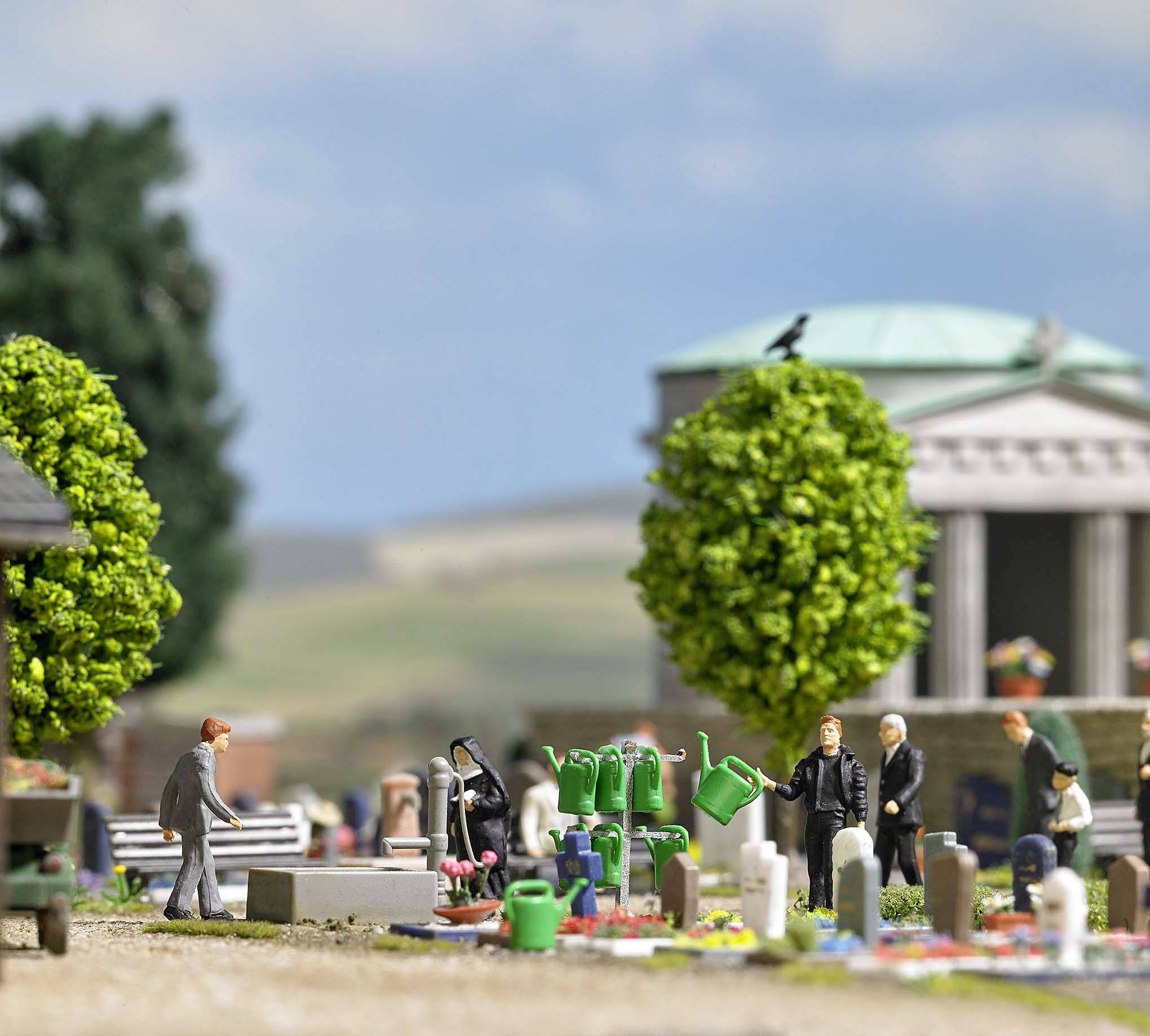 Personen op een miniatuurkerkhof met grafstenen, bomen en gieterjes. Op de achtergrond zijn een tempelgebouw en landschap zichtbaar.