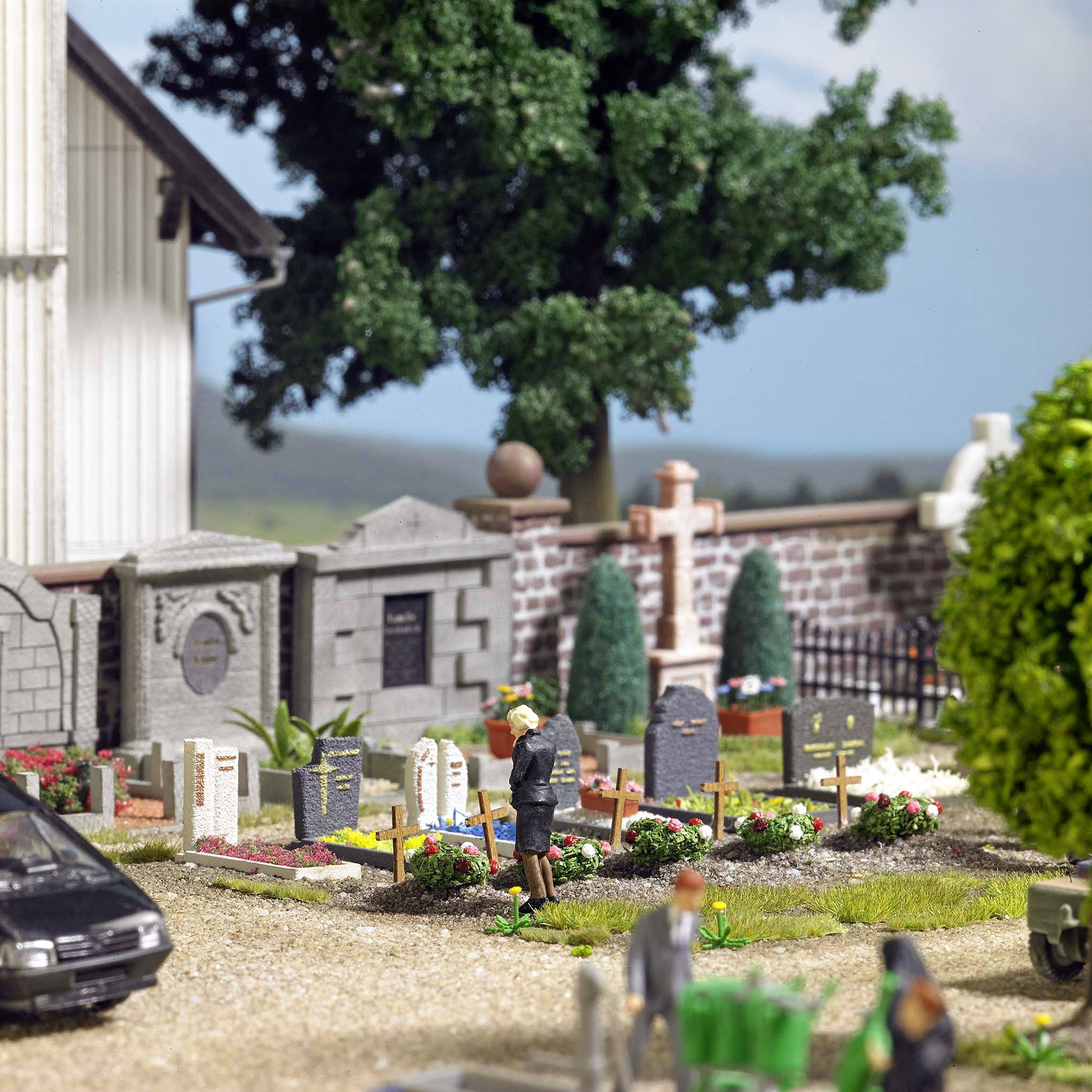 Een miniatuurkerkhof met grafstenen, een biddende figuur, bomen en een auto op de voorgrond, omgeven door een groene landschap en blauwe lucht.