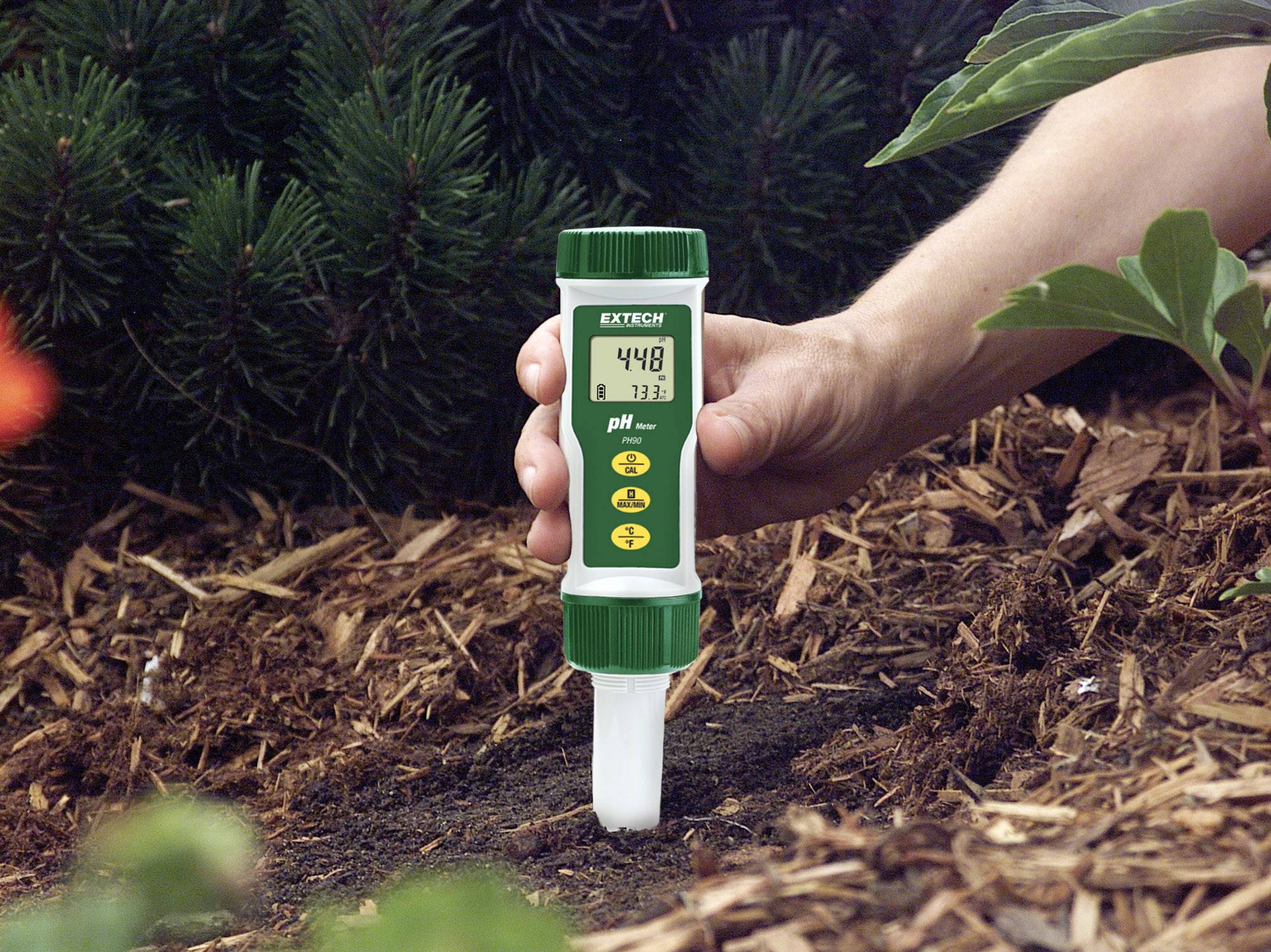 Een hand meet de pH-waarde van de bodem met een digitaal pH-meetapparaat dat een waarde van 4,48 aangeeft. Omgeving: tuingrond, planten.