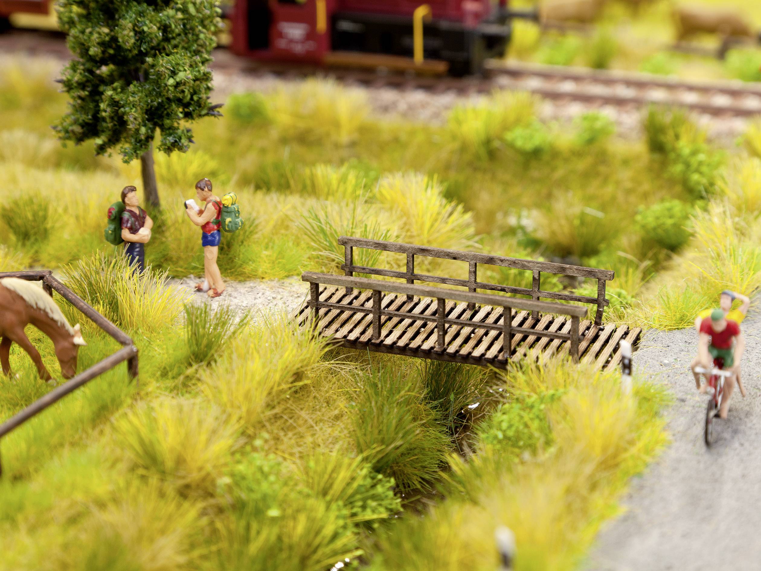 Een miniatuurscène toont wandelaars op een pad langs een brug, terwijl een andere figuur op een fiets rijdt. Op de achtergrond zijn sporen te zien.