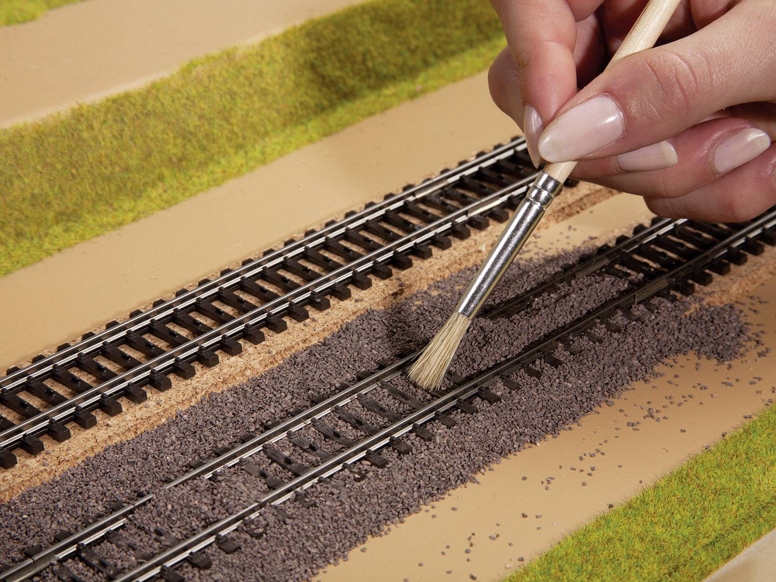 'Een hand schildert fijne steentjes op een modelspoorrailtraject om een realistische landschap te creëren.'