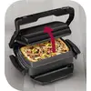 Een contactgrill die een pizza met pepperoni en olijven bakt. Een rode pijl wijst naar de handgreep van de grill, die opengaat.