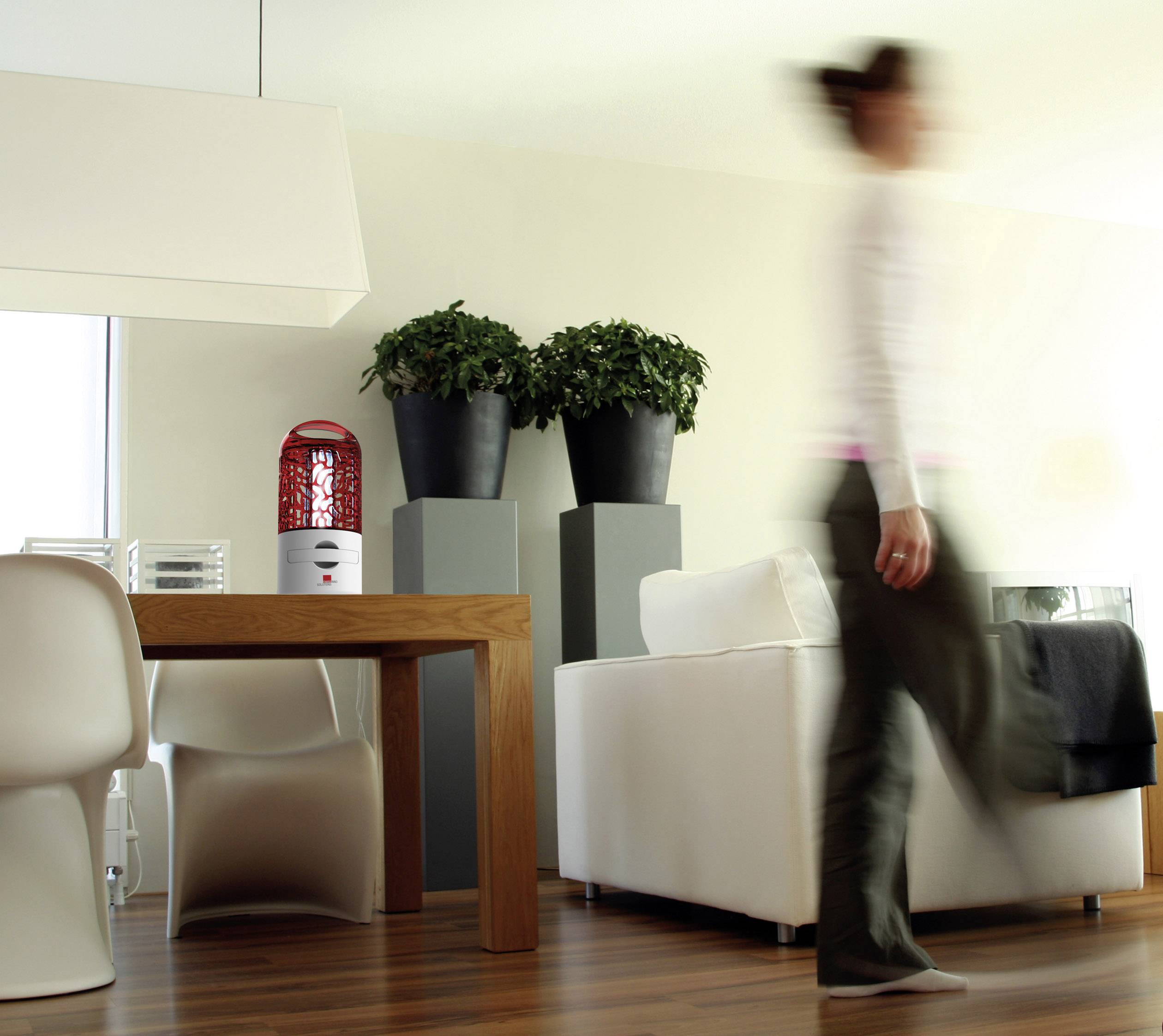 Een persoon loopt langs een modern ingerichte ruimte met een houten tafel en witte stoelen; op de achtergrond planten op sokkels.