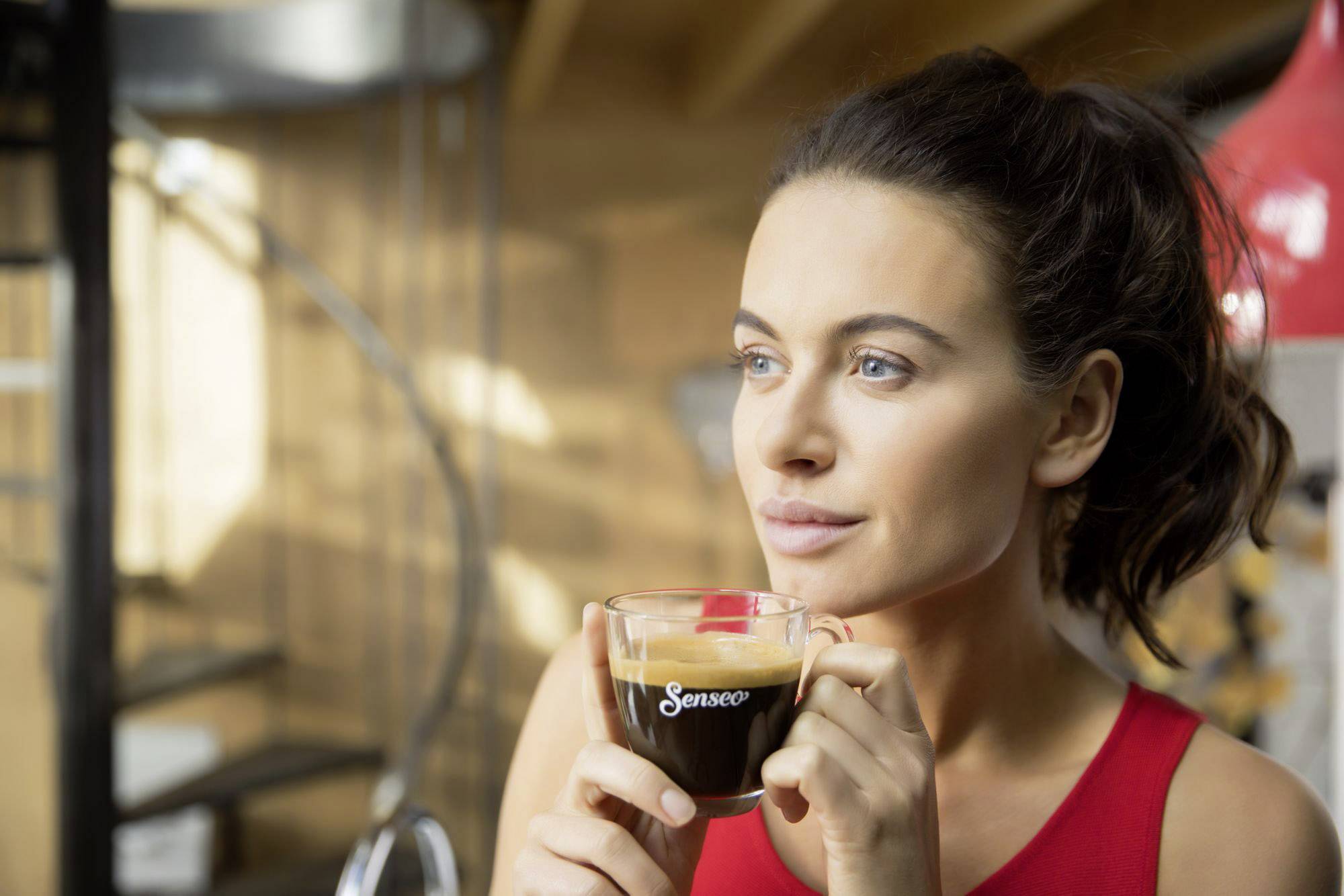 Een vrouw houdt een kopje koffie vast en kijkt peinzend opzij. Op de achtergrond is een trap.