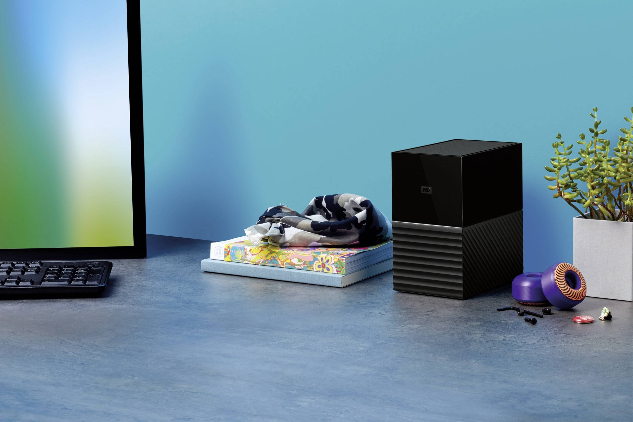 Op een bureau staan een computerscherm, een toetsenbord en een externe harde schijf. Ernaast liggen boeken en planten.