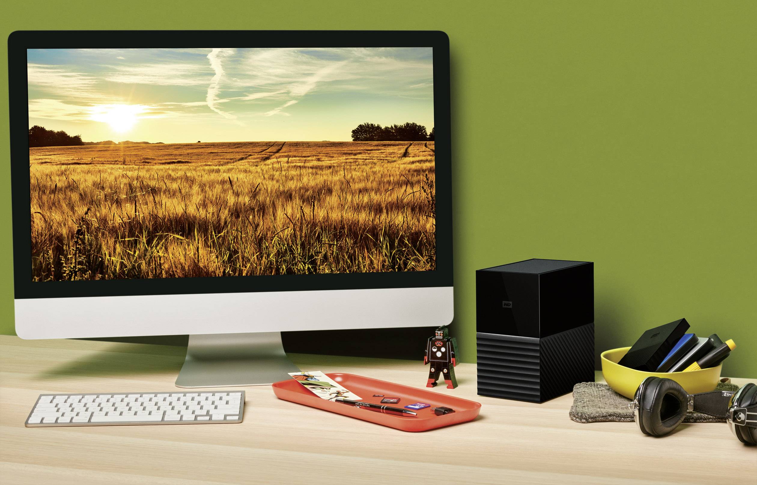 Een computerscherm op een bureau toont een landschap met een geel tarweveld bij zonsondergang. Naast het scherm liggen bureaubenodigdheden.