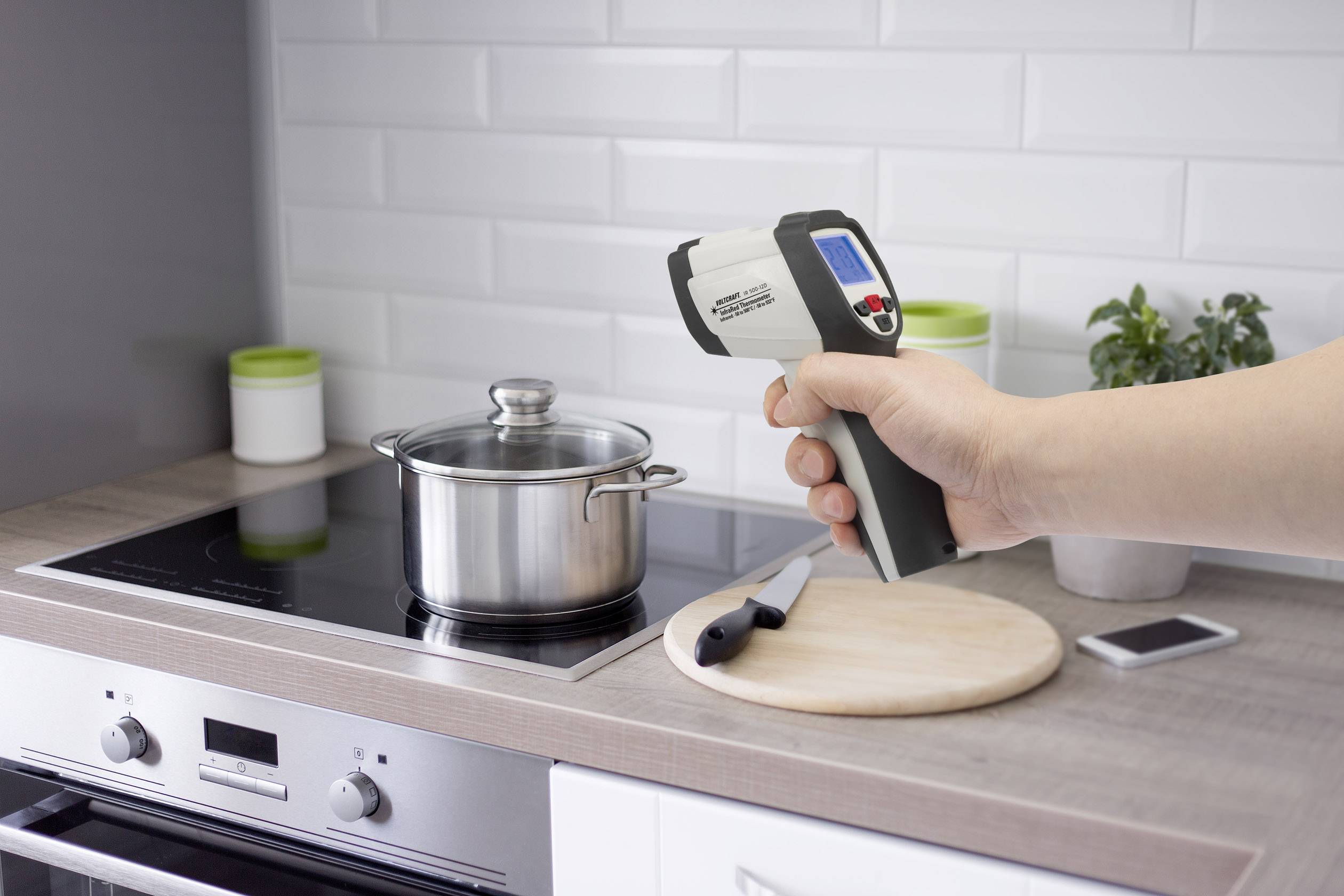 Een hand houdt een infrarood-thermometer vast die gericht is op een pan op het fornuis. De meetwaarde wordt weergegeven op het digitale display.