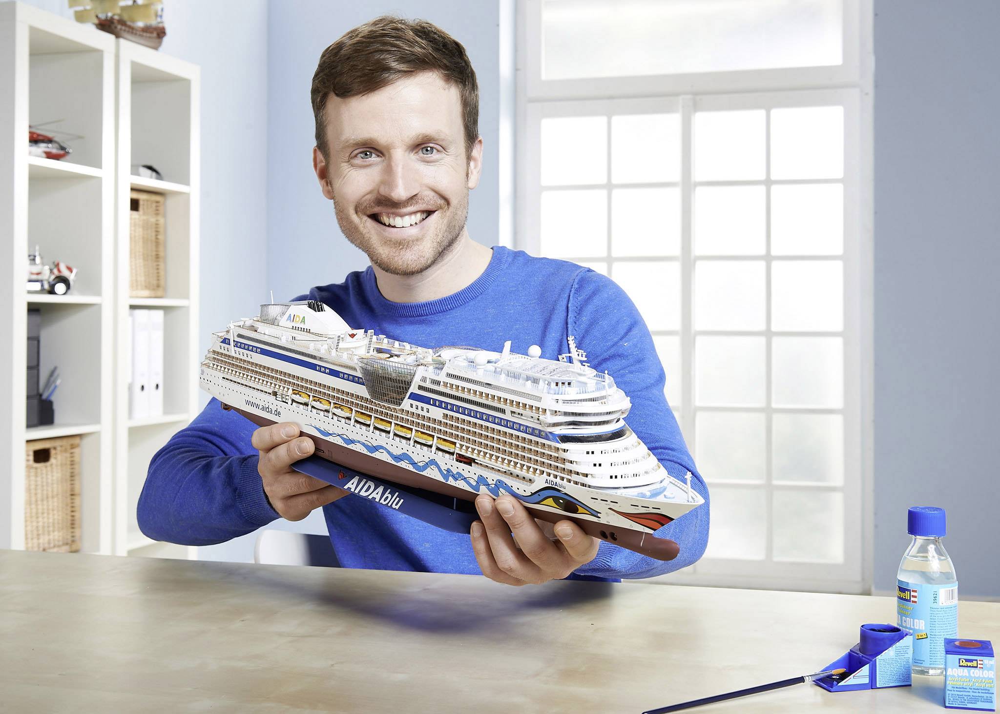 Een man houdt een model van een cruiseschip met het opschrift 'AIDAblu' in een lichte ruimte. Op de achtergrond een raam en planken.