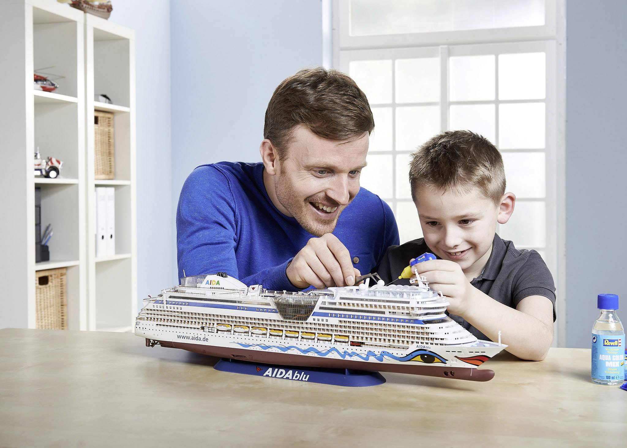 Een man en een jongen bouwen samen een modelschip van de 'AIDAblu' op een tafel. Op de achtergrond zijn planken en een raam.