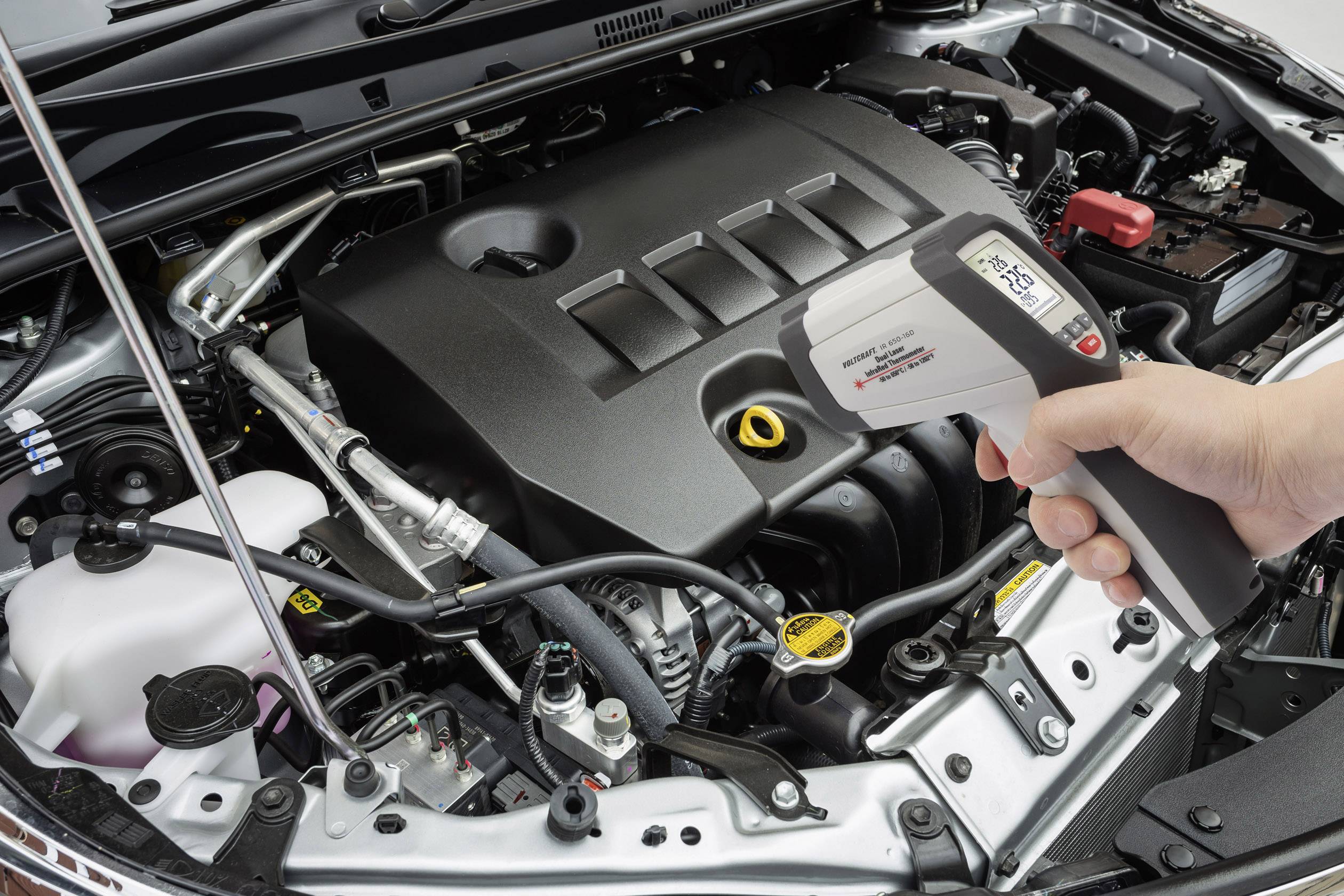 Een automonteur meet met een infrarood-thermometer de temperatuur van een motor die zich in een open motorruimte van een auto bevindt.