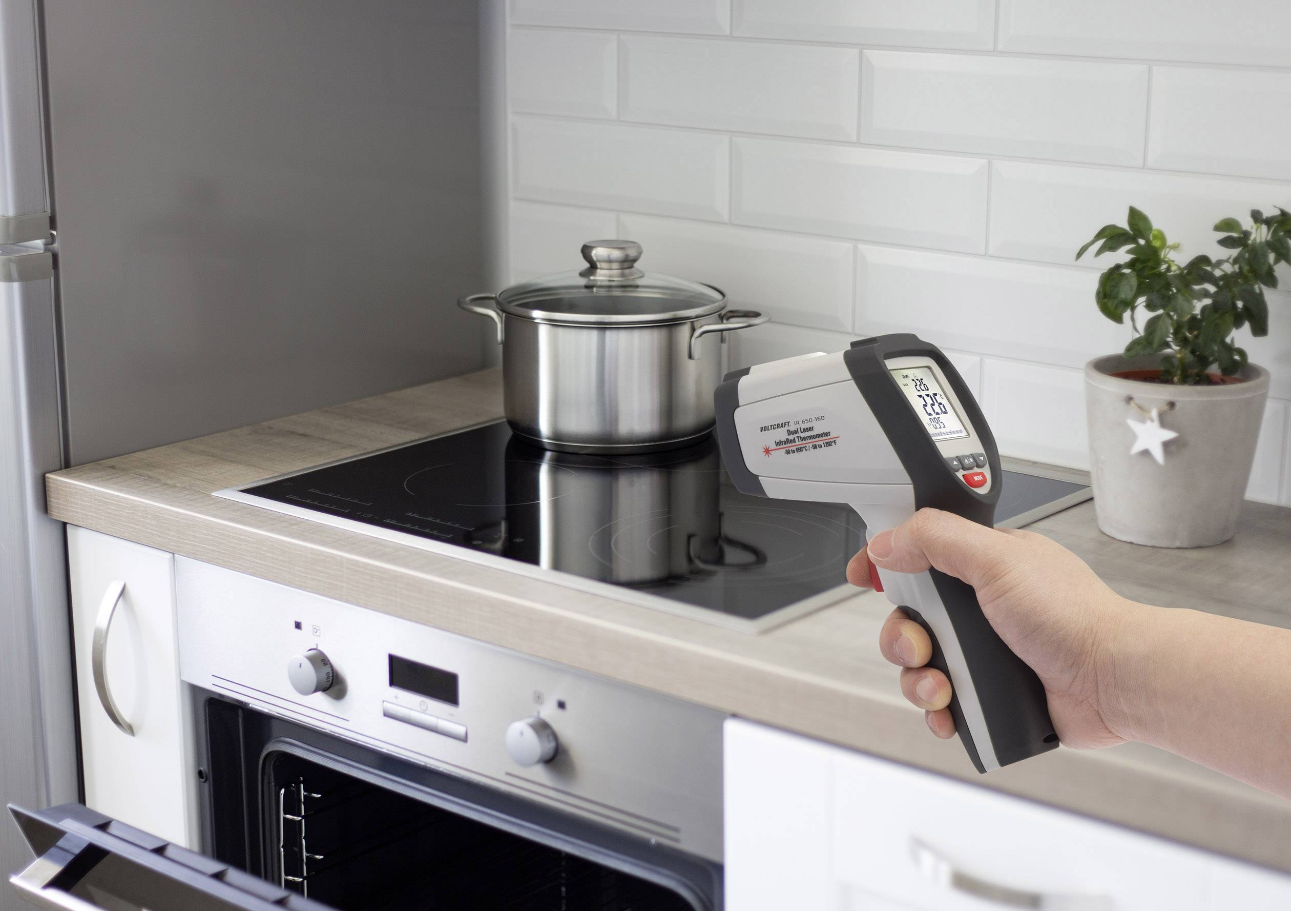 Een hand houdt een infraroodthermometer vast en meet de temperatuur van een pan op een inductiekookplaat in een moderne keuken.