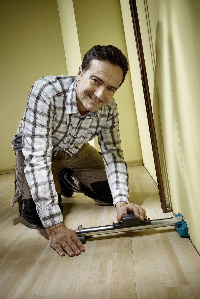 Een man knielt op een houten vloer en gebruikt een gereedschap om de muur op te meten.