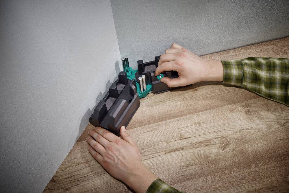 Twee handen monteren een pluggenset in een hoek van een kamer op een houten tafel. Muur en tafel staan loodrecht op elkaar.