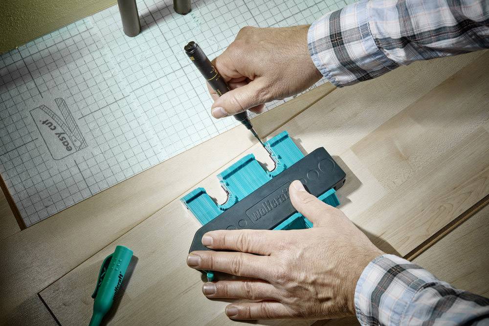 Een persoon houdt een turquoise gereedschapsdeel vast en gebruikt een pen om te markeren op hout, op een tafel met geruit papier.