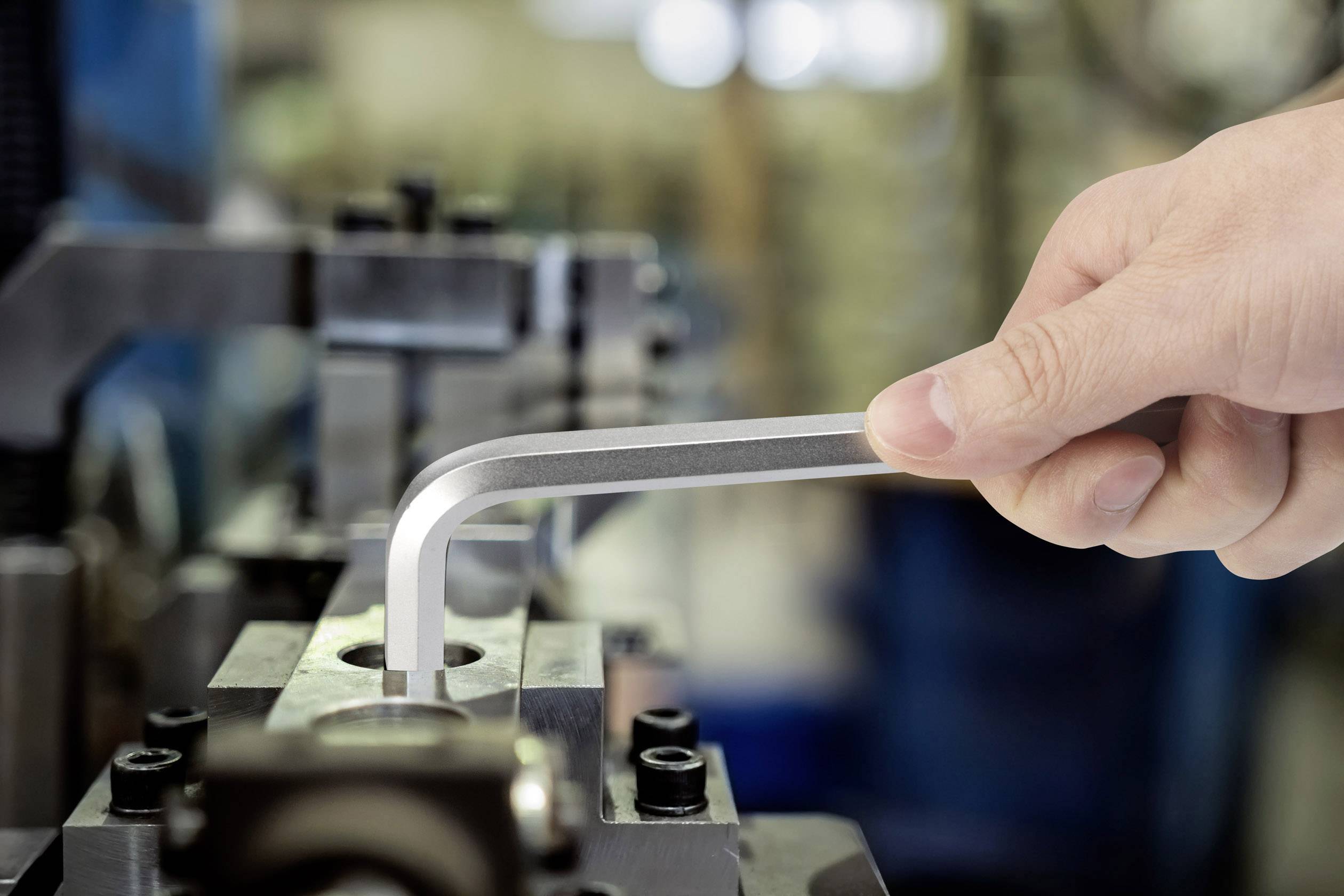 Een hand gebruikt een inbussleutel om een machine af te stellen in een werkplaatsomgeving. Op de achtergrond zijn onscherpe apparaten zichtbaar.