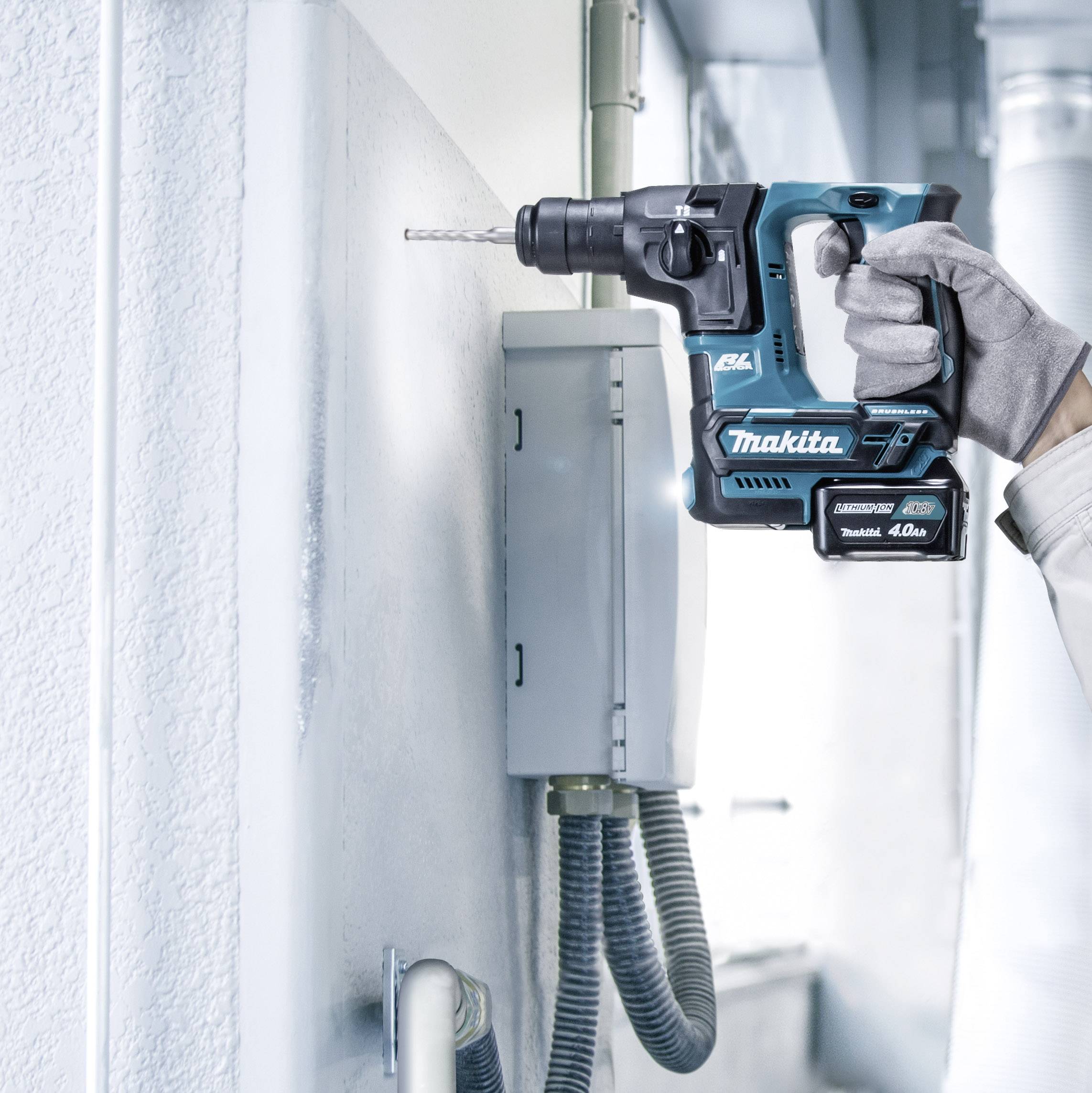 'Een persoon houdt een Makita-boormachine en boort in een grijze muur. De boormachine is blauw en wordt gebruikt in een industriële omgeving.'