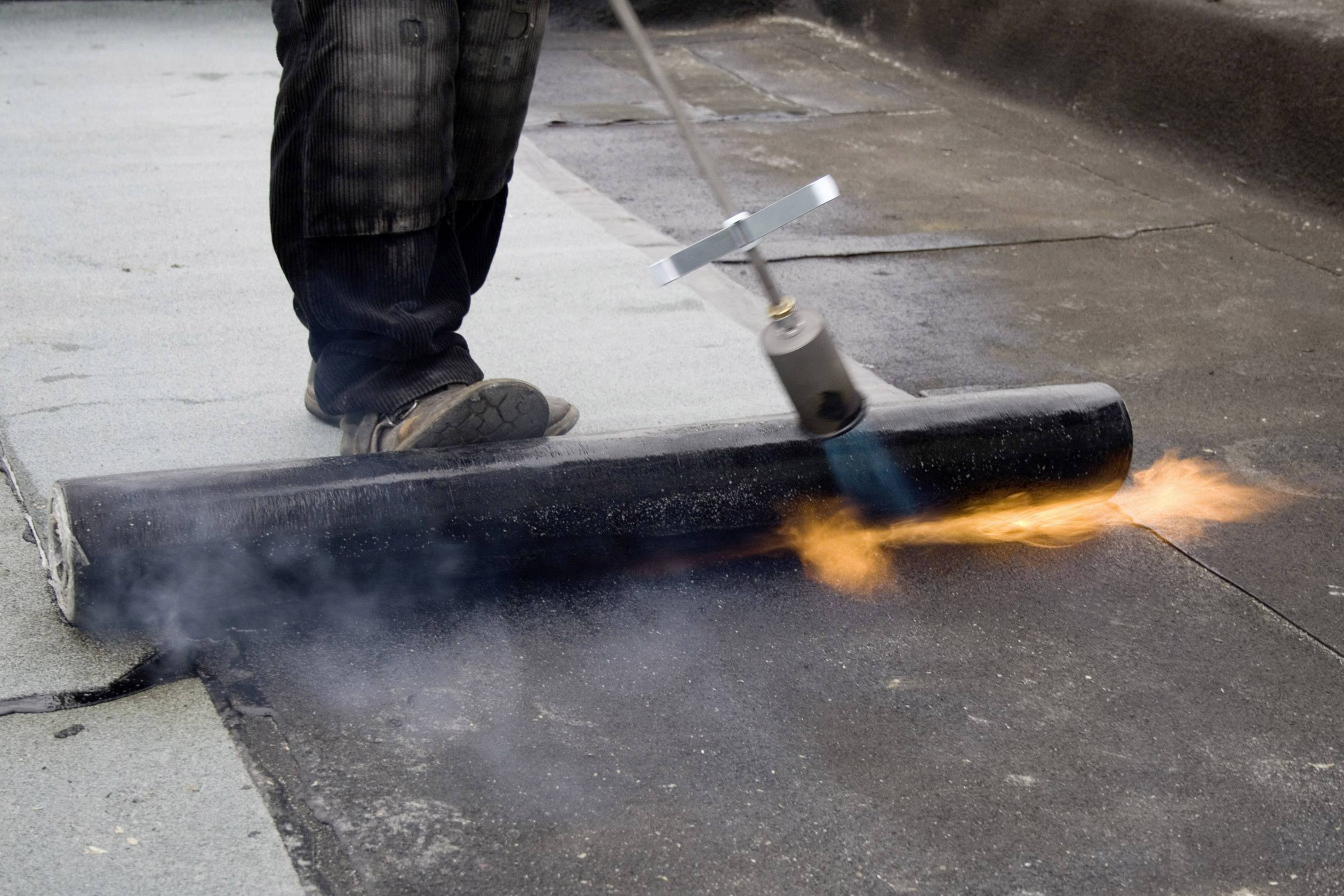 Een persoon gebruikt een vlam om dakmateriaal op een oppervlakte te bevestigen. De rol van zwart materiaal wordt verhit.