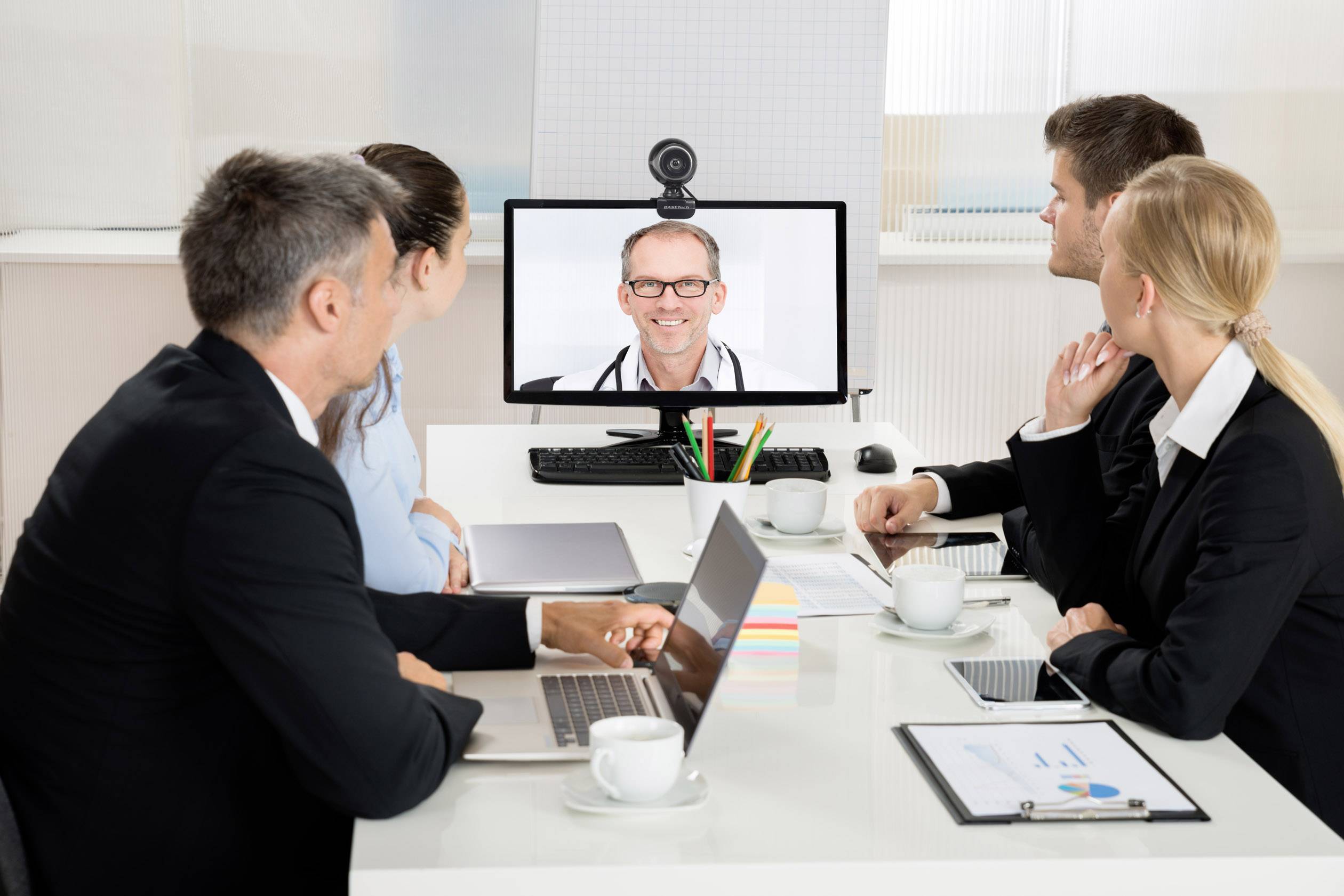 Vier personen in een kantoor kijken naar een computerscherm waarop een arts te zien is in een videogesprek.