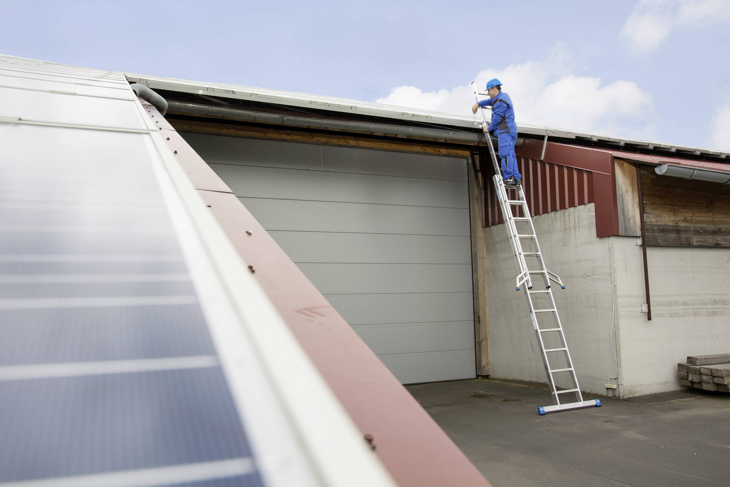 Een arbeider in een blauwe overall staat op een ladder en repareert het dak van een magazijn. Links zijn zonnepanelen zichtbaar.