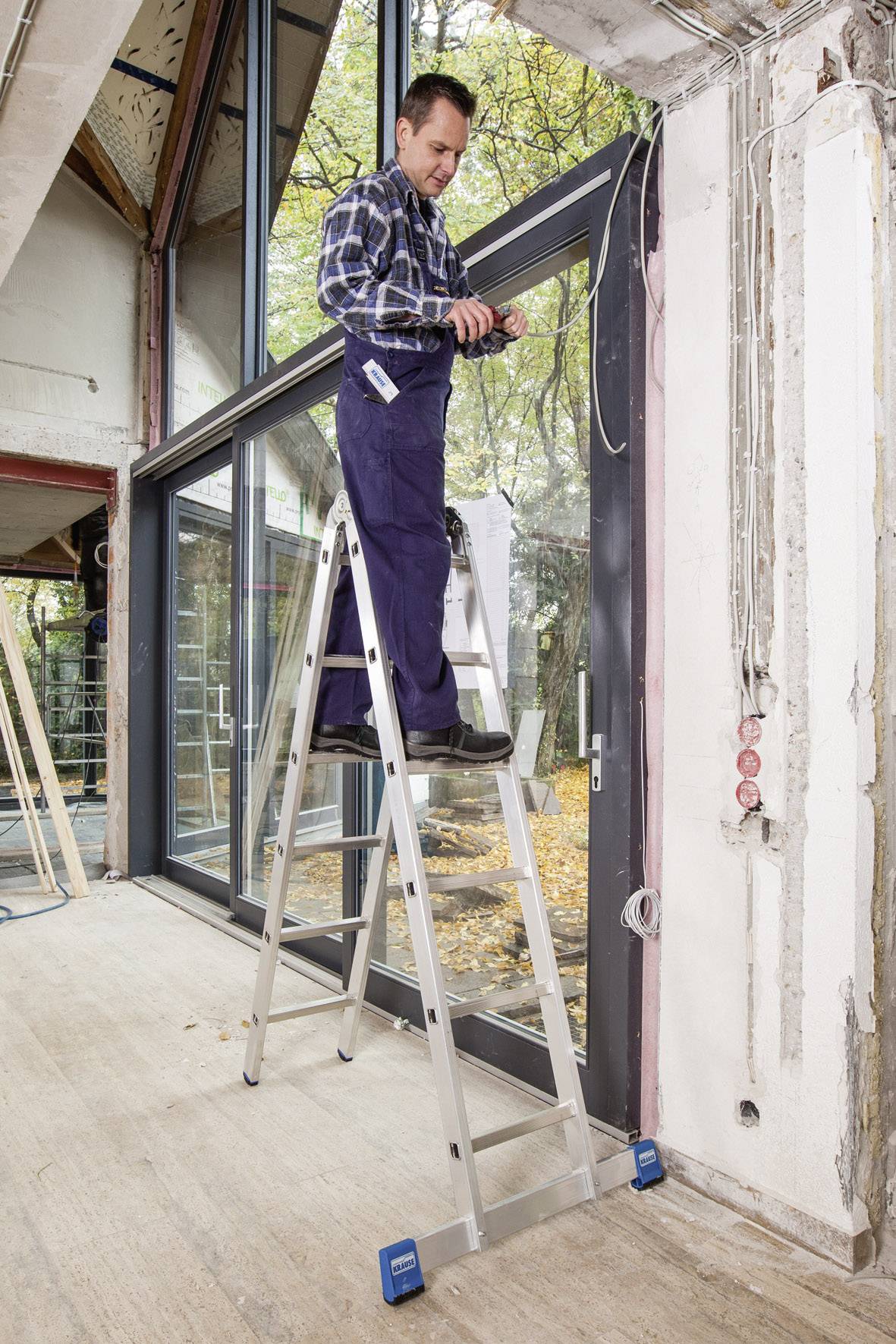 Een persoon staat op een ladder in een ruimte met grote ramen en werkt aan elektrische kabels aan een muur.