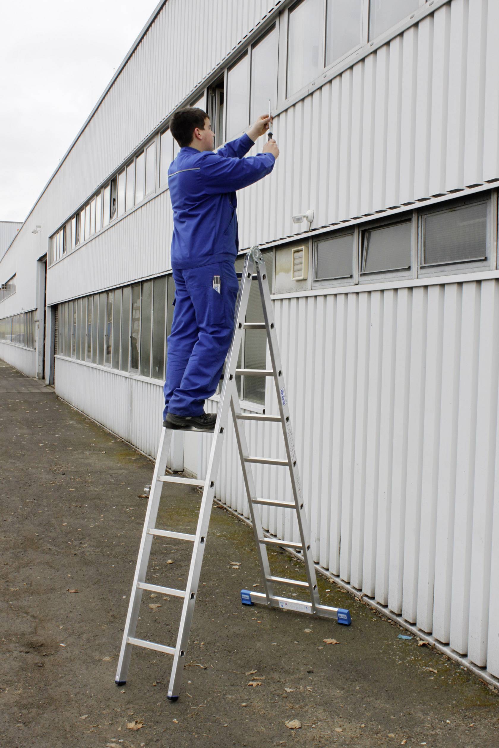 Een man in blauwe werkkleding staat op een ladder en werkt aan de buitenmuur van een industrieel gebouw.