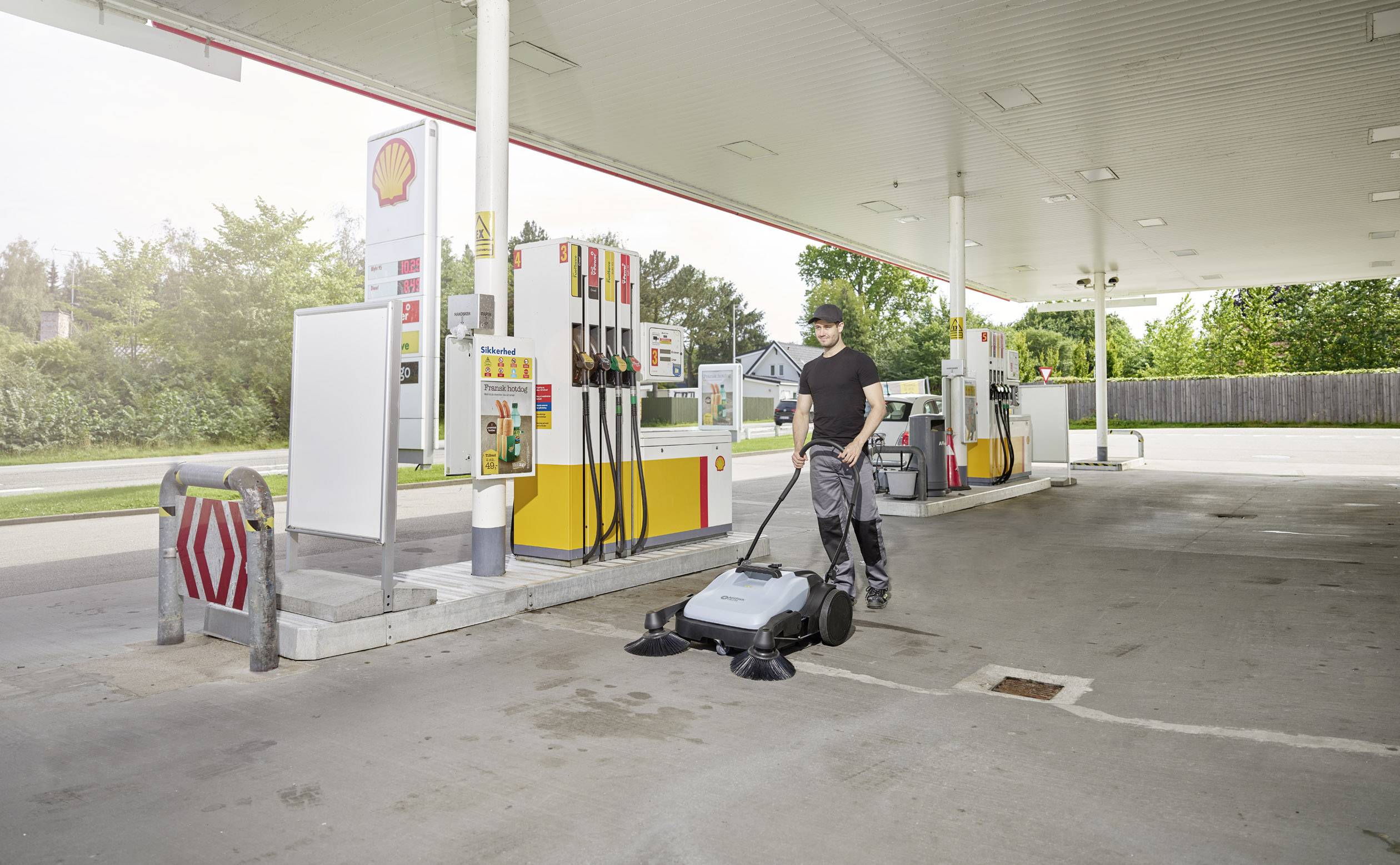 Een persoon reinigt de grond van een tankstation met een veegmachine. Op de achtergrond zijn pompinstallaties en bomen zichtbaar.