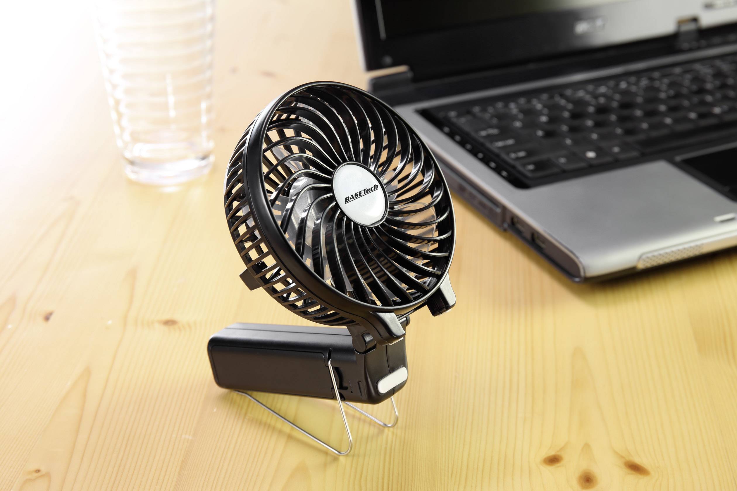 Een kleine, zwarte tafelventilator staat aangeschakeld naast een laptop op een houten tafel. Ernaast staat een leeg glas.