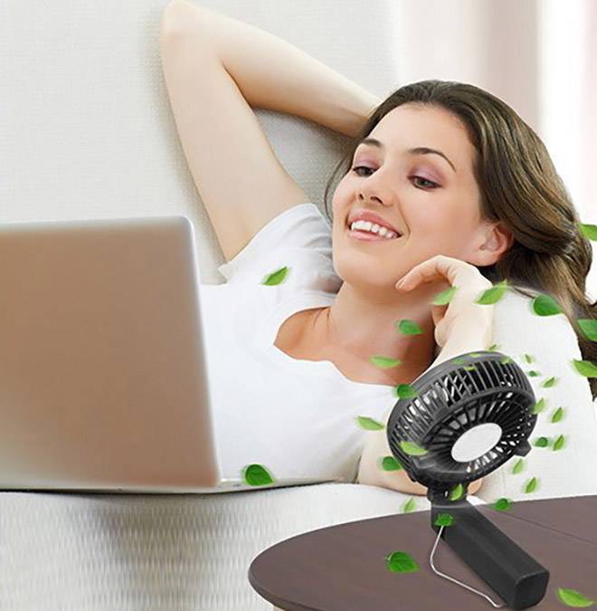 Een vrouw ontspant op een bank met een laptop, terwijl een tafelventilator groene bladeren naar haar toe waait, wat een verfrissende bries suggereert.