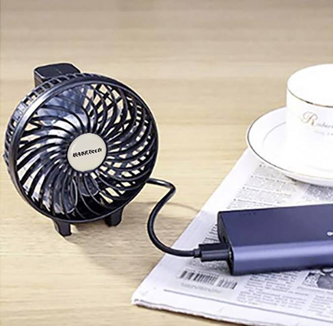 Een kleine, zwarte USB-ventilator naast een koffiemok op een tafel die bedekt is met een krant. De ventilator is aangesloten op een powerbank.