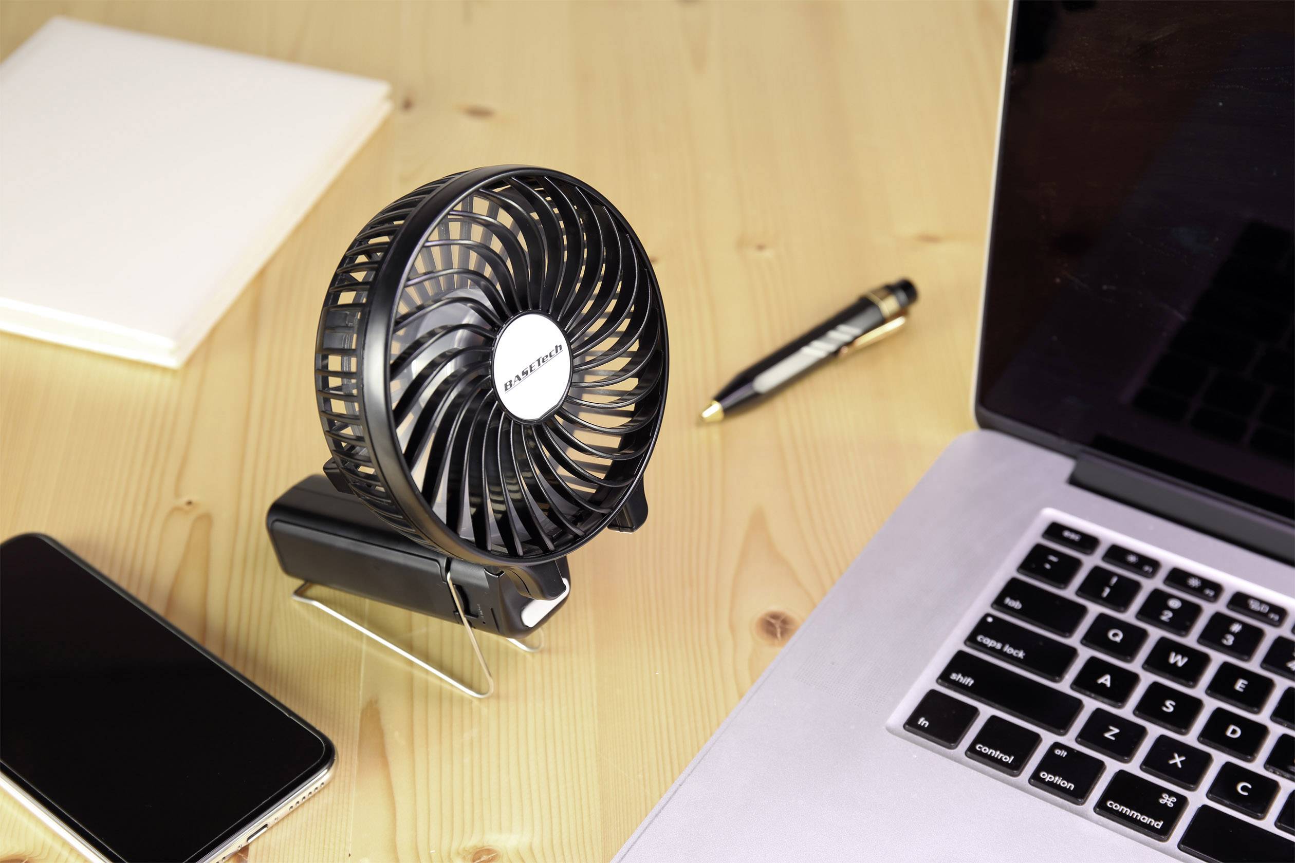 Een kleine ventilator op een bureau naast een laptop, een smartphone, een notitieboekje en een pen.