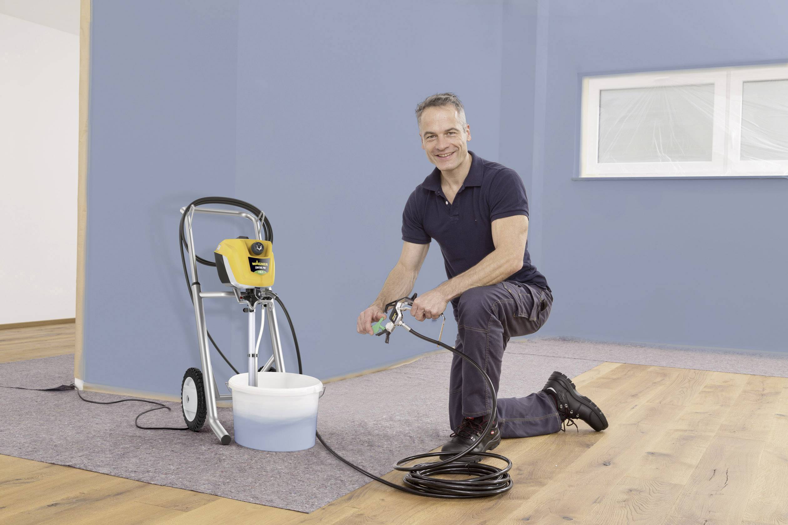 Een man knielt op een houten vloer naast een verfmachine. Hij houdt een verfspuitopzetstuk vast en bereidt zich voor op schilderwerkzaamheden.