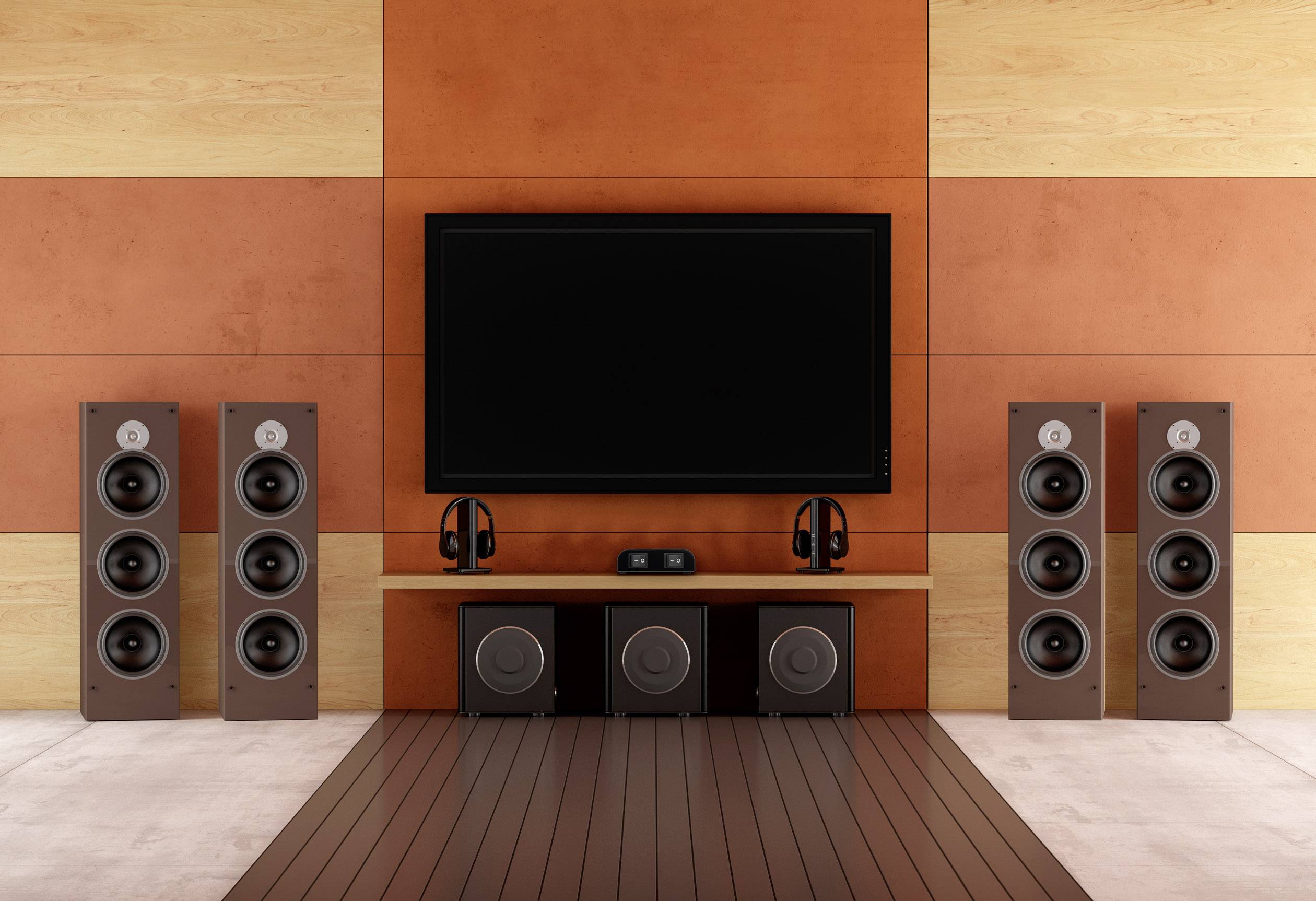 Thuisbioscoop-setup met een groot flatscreen-tv-toestel, vier vloerstaande luidsprekers en drie subwoofers voor een oranje-bruine muur.