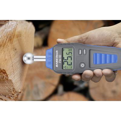 Een vochtmeter toont '20%' aan, terwijl deze tegen hout wordt gehouden. Op de achtergrond zijn boomstammen zichtbaar.