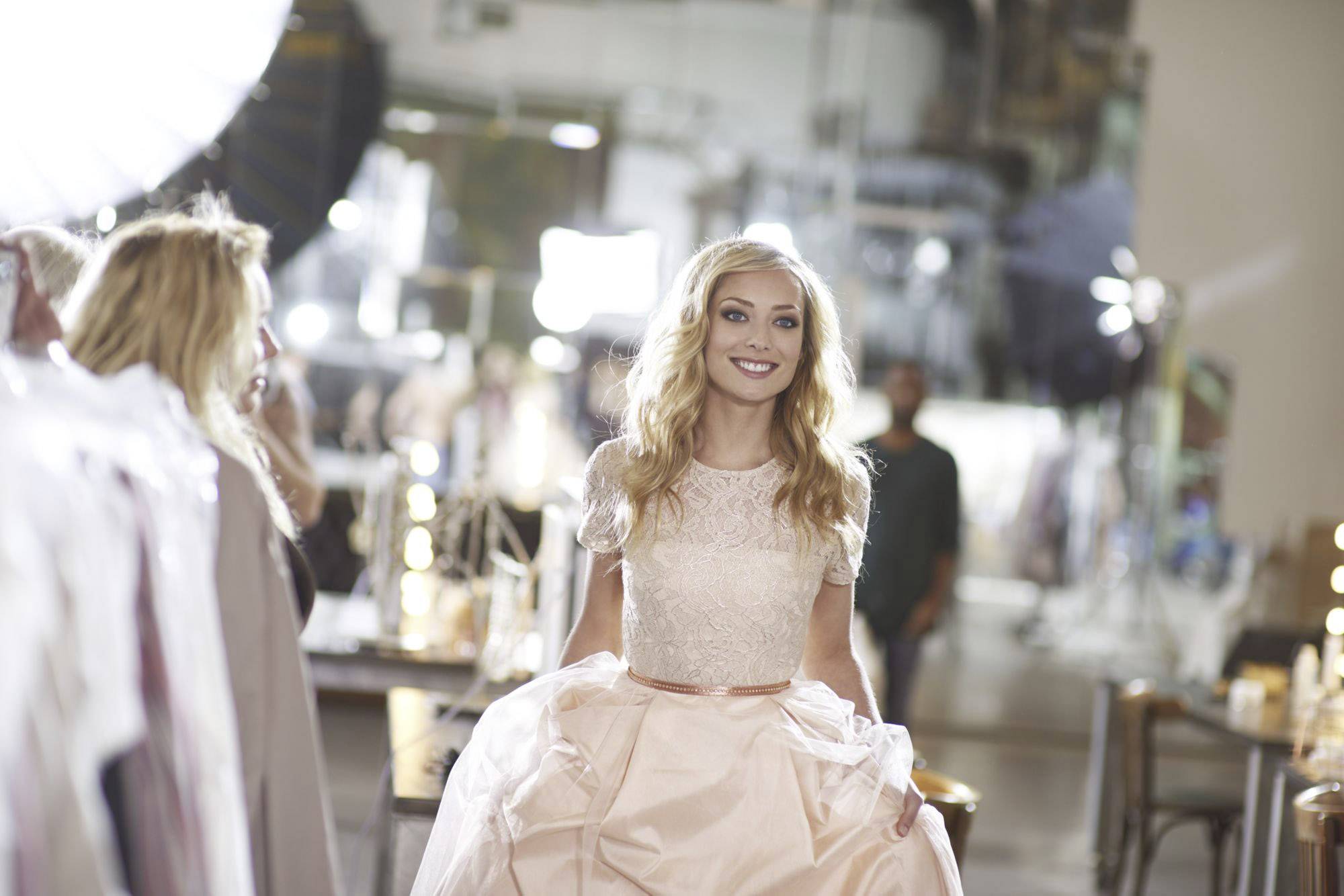 Een glimlachende vrouw in een elegant, roze jurk tijdens een fotoshoot, met belichting- en camera-apparatuur op de achtergrond.
