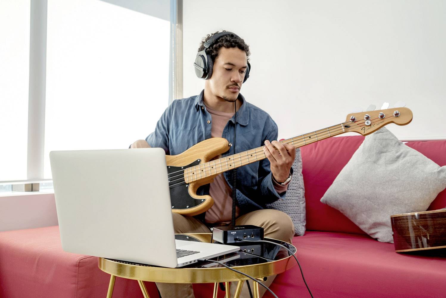 Een man speelt basgitaar op een rode bank, draagt koptelefoons en kijkt naar een laptop op een tafel voor hem.