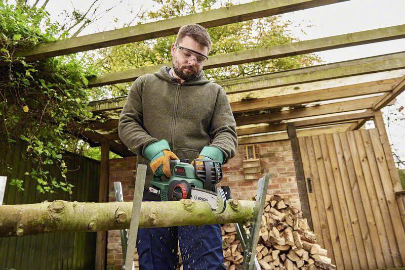 Een man met veiligheidsbril en handschoenen gebruikt een kettingzaag om een dikke tak door te zagen op een zaagbok in de tuin.