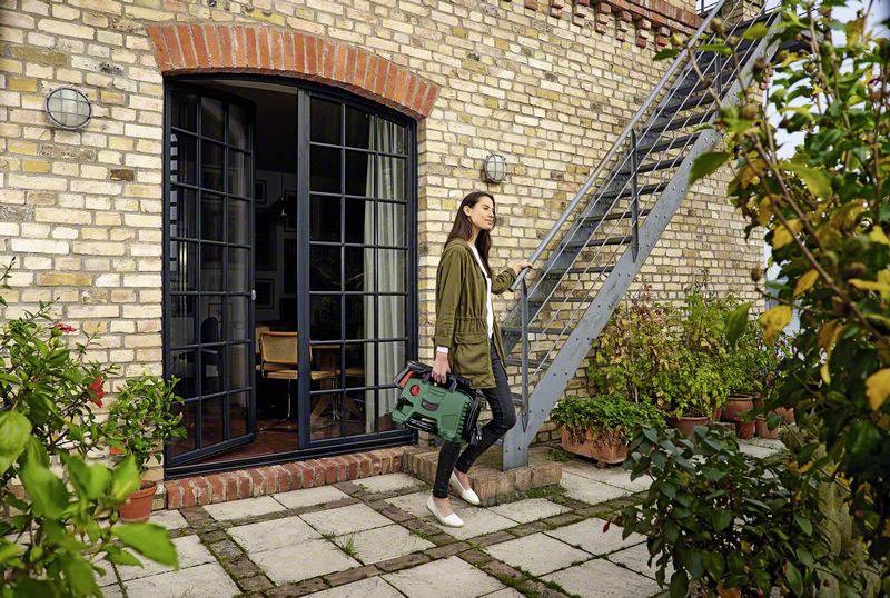 Een vrouw staat met een tuinschaar in de hand op een terras voor een bakstenen huis met glazen deuren en planten.