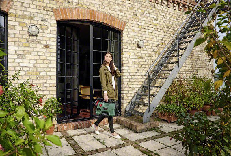 Vrouw in casual kleding draagt een groene gereedschapstas en staat op een terras voor een bakstenen huis met een metalen trap.