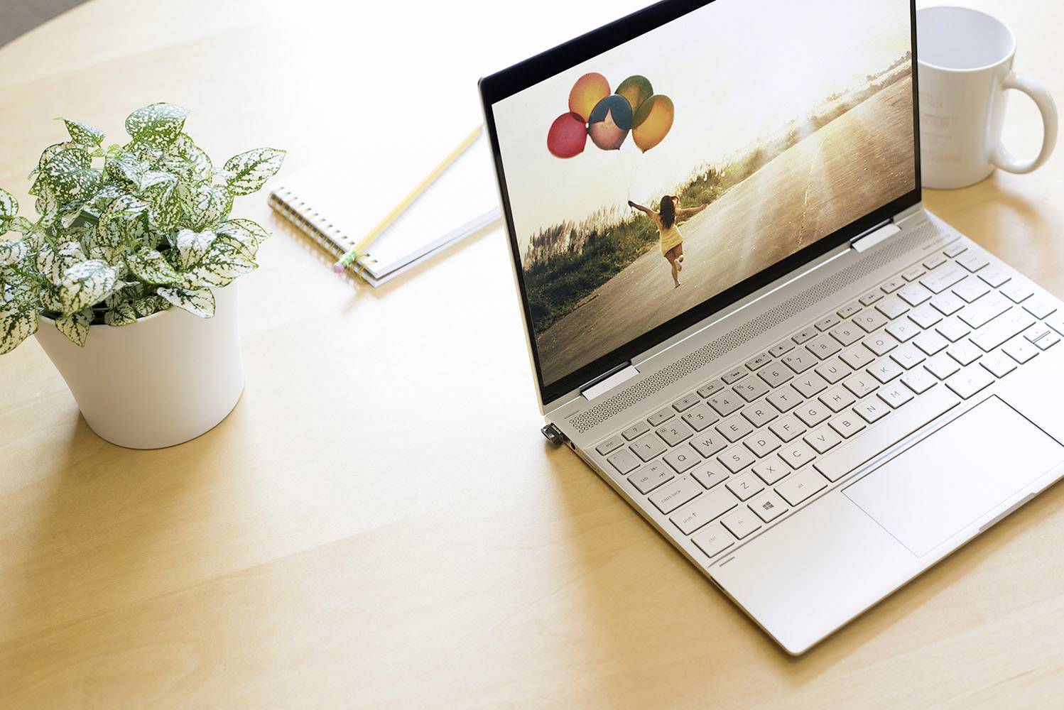 Zilveren laptop op een houten tafel met een foto van een kind dat ballonnen vasthoudt. Ernaast een koffiekopje, een plant en een notitieboekje.